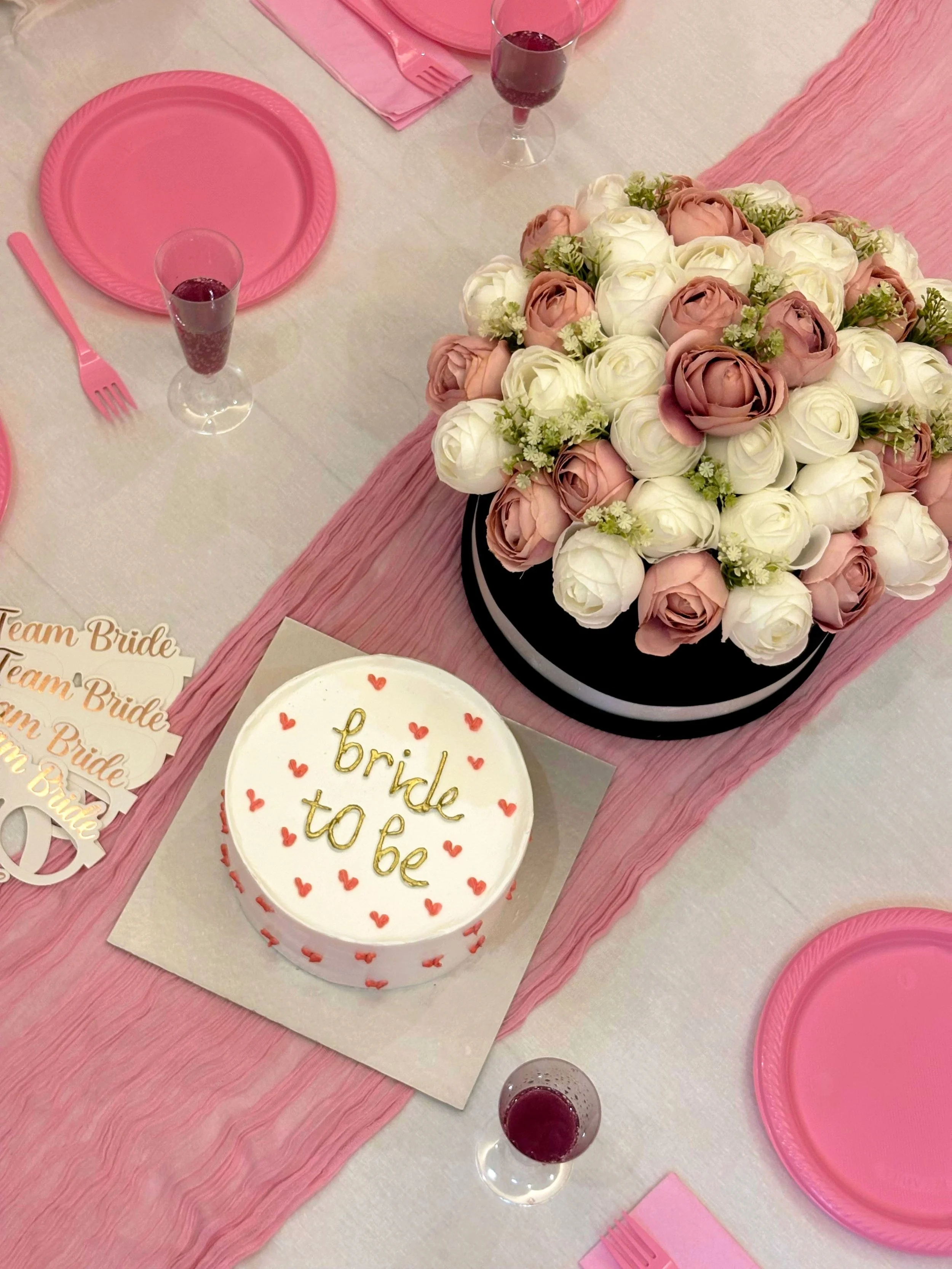 The image shows a pink-themed wedding or bridal shower table setting with a bouquet of white and pink roses, pink plates, pink napkins, pink utensils, wine glasses with red drink, and a white cake with red hearts and gold writing that says "bride to be."