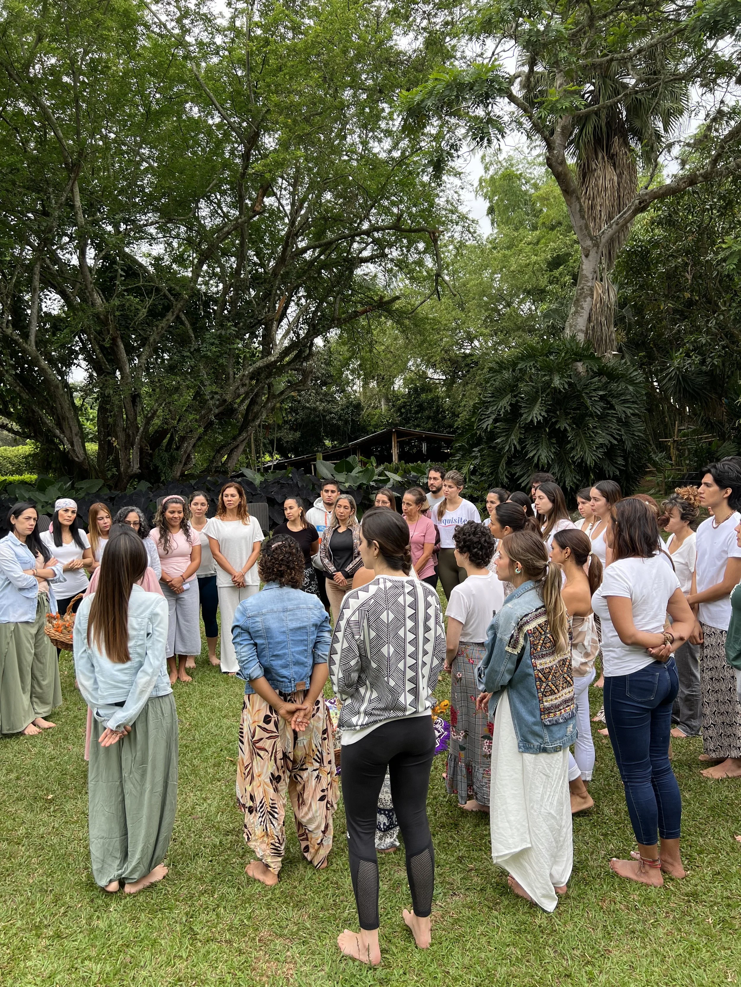 People participating in a group outdoor yoga session on colorful mats and bean bags in a park-like setting with trees, rocks, and stone structures.