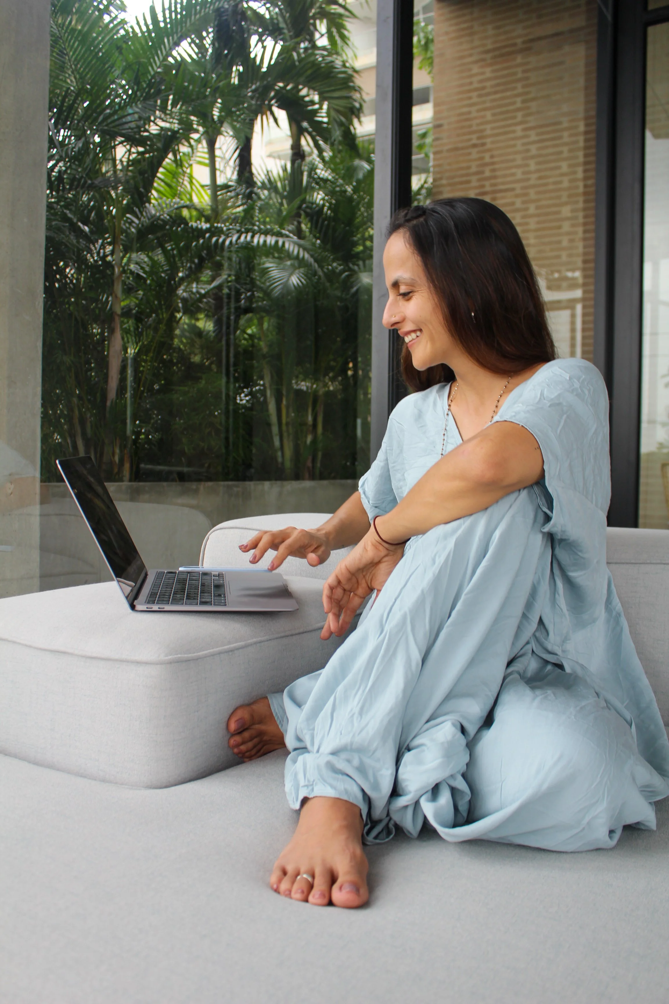Woman sitting on a couch, smiling, while using a laptop, with a view of greenery outside the window.