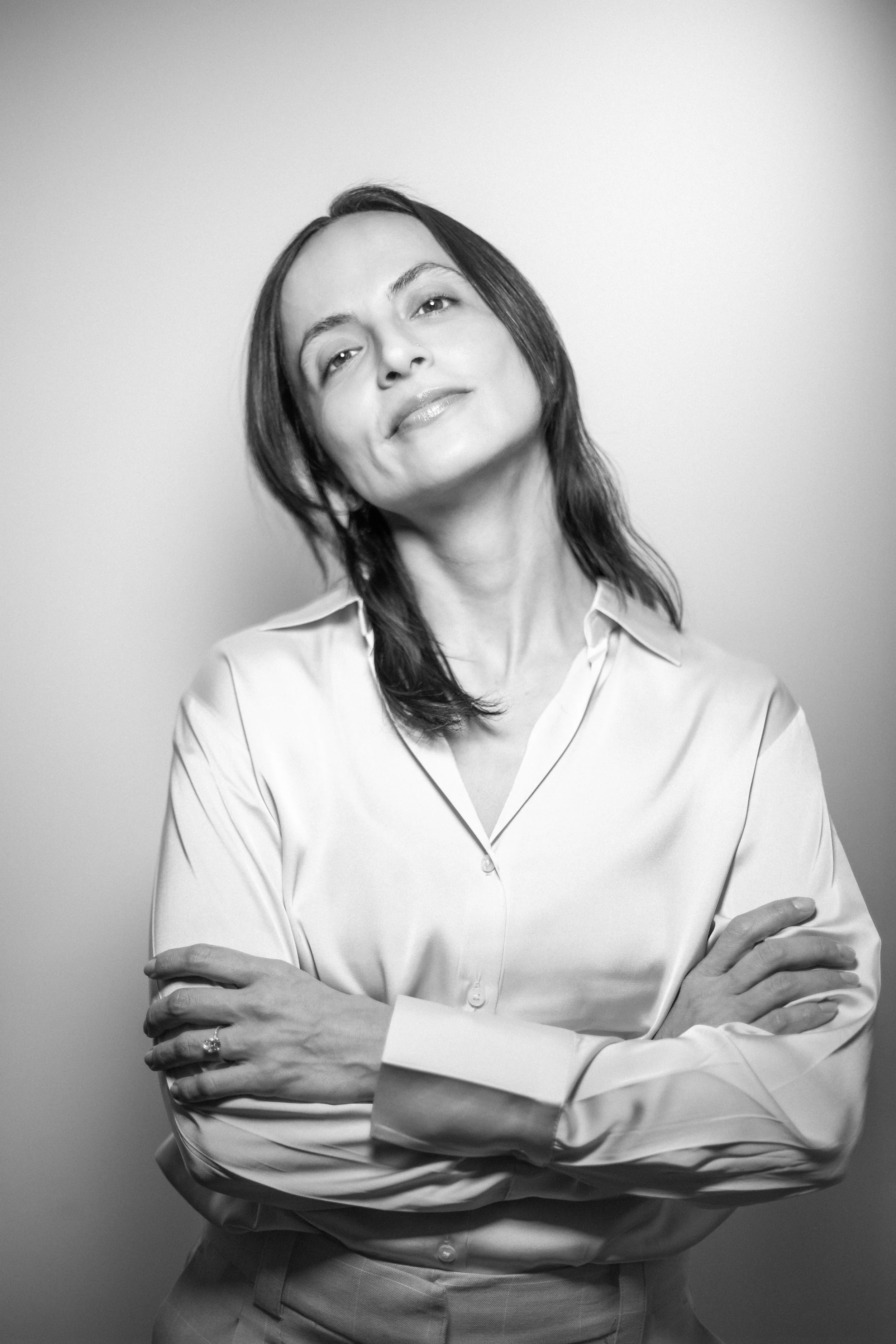 Black and white portrait of a woman with shoulder-length dark hair, crossing her arms and wearing a button-down shirt, standing against a plain background.