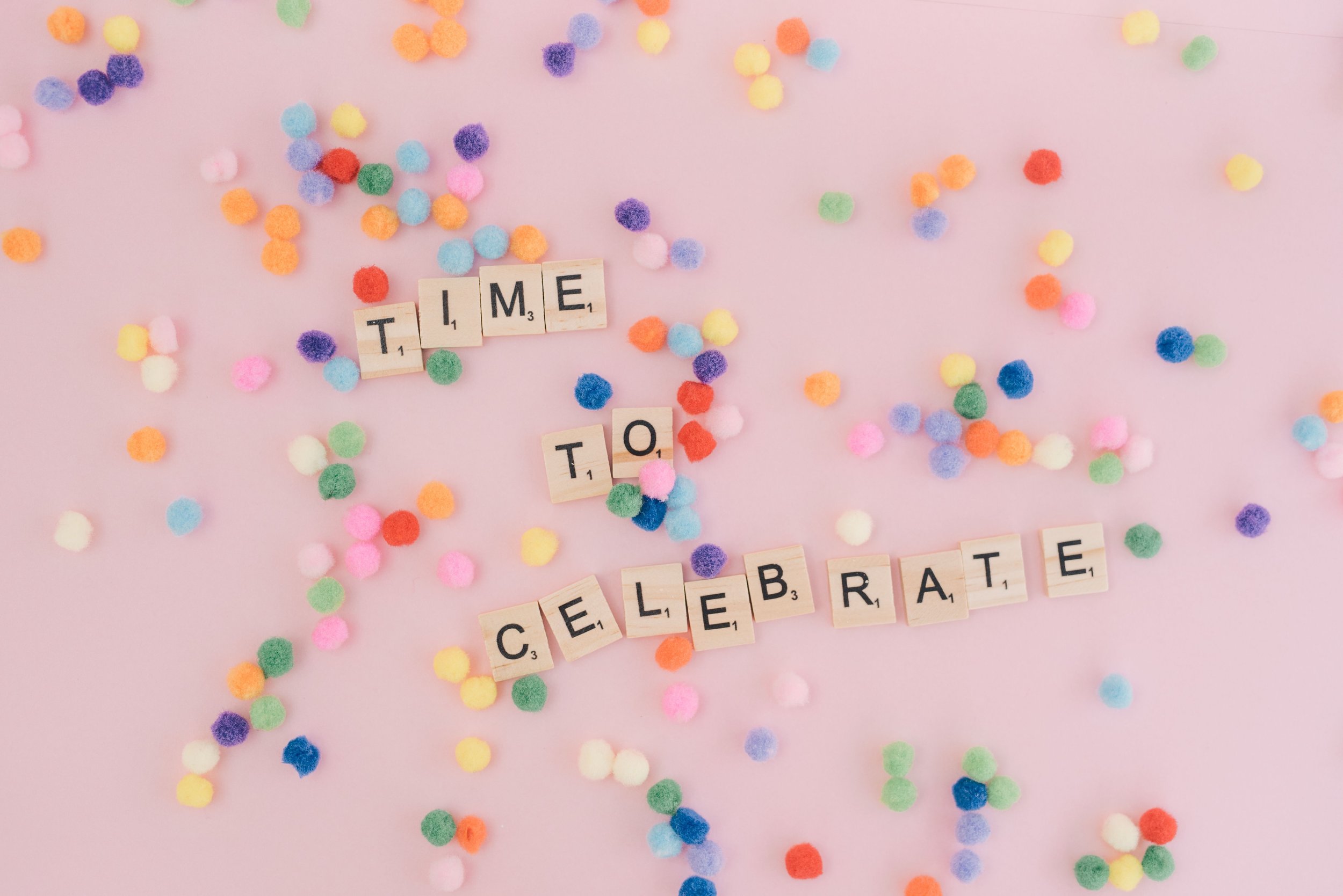 Colorful pom-poms scattered on a pink background with scrabble tiles spelling out 'TIME TO CELEBRATE'.
