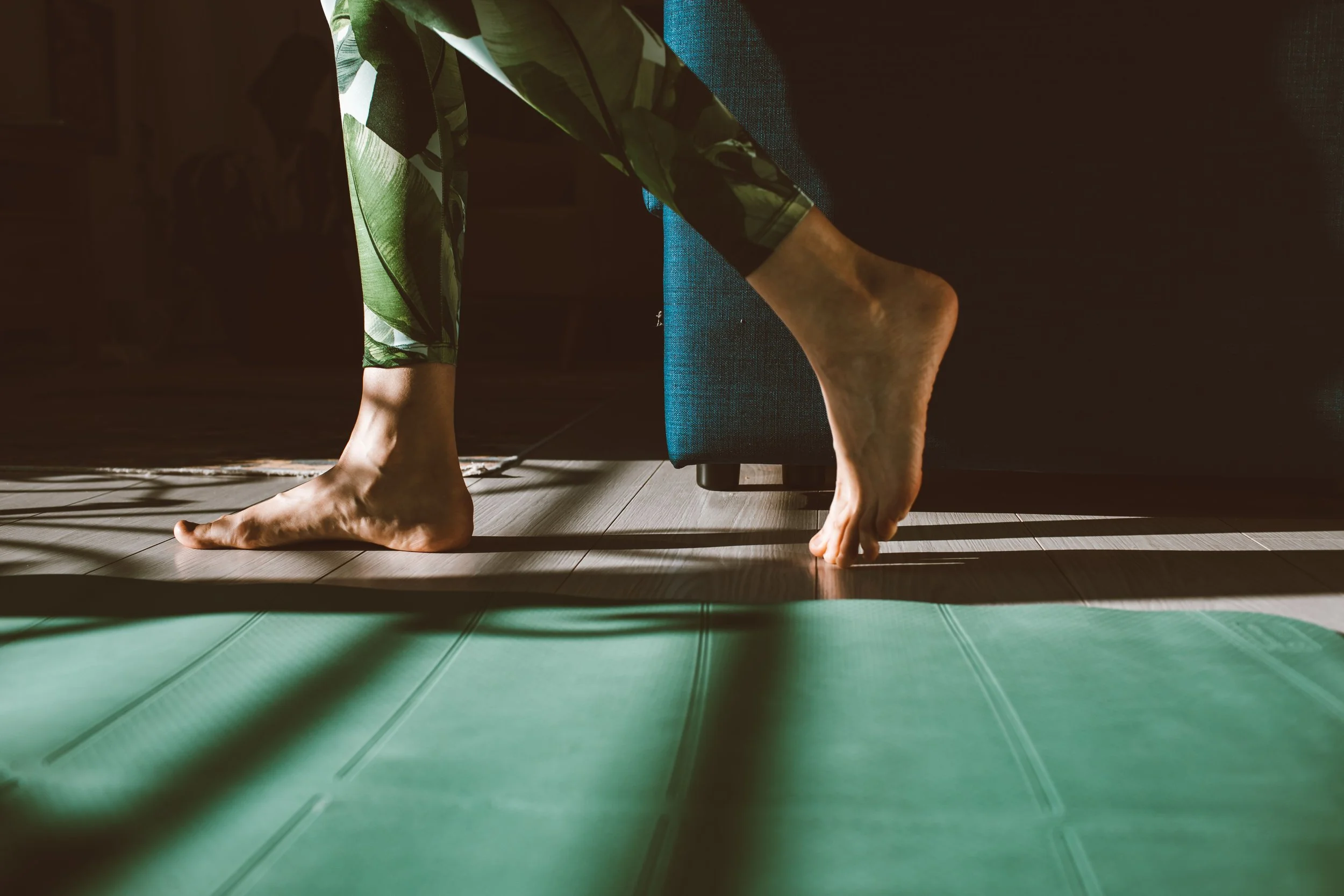Close-up of a person in green and white patterned leggings practicing yoga or stretching on a green yoga mat, with their feet on the floor near a dark couch.