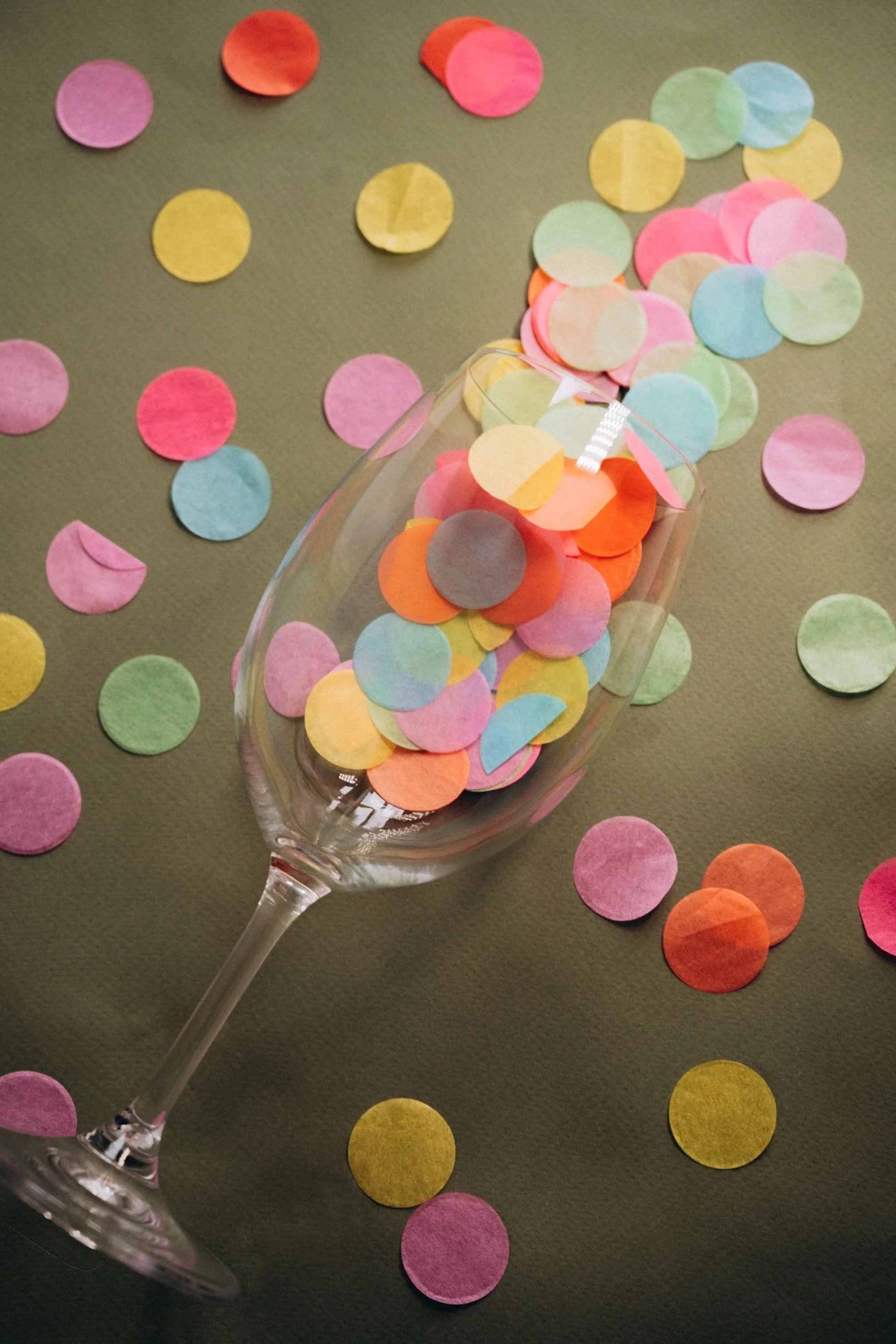 A wine glass filled with colorful paper confetti circles is resting on a dark surface, with similar confetti scattered around.