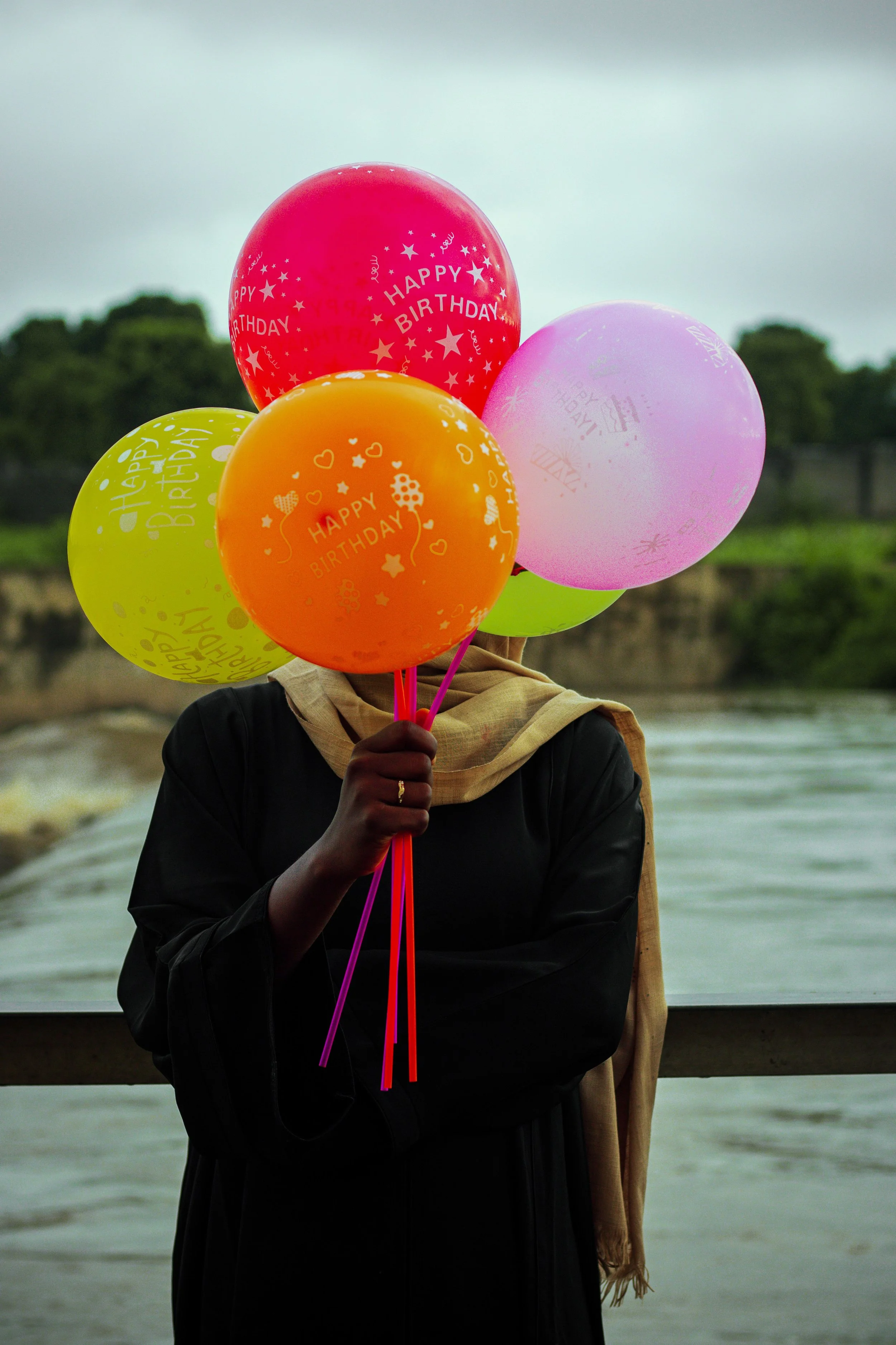 Person holding multiple colorful balloons with 'Happy Birthday' text printed on them, standing near water with trees in the background.