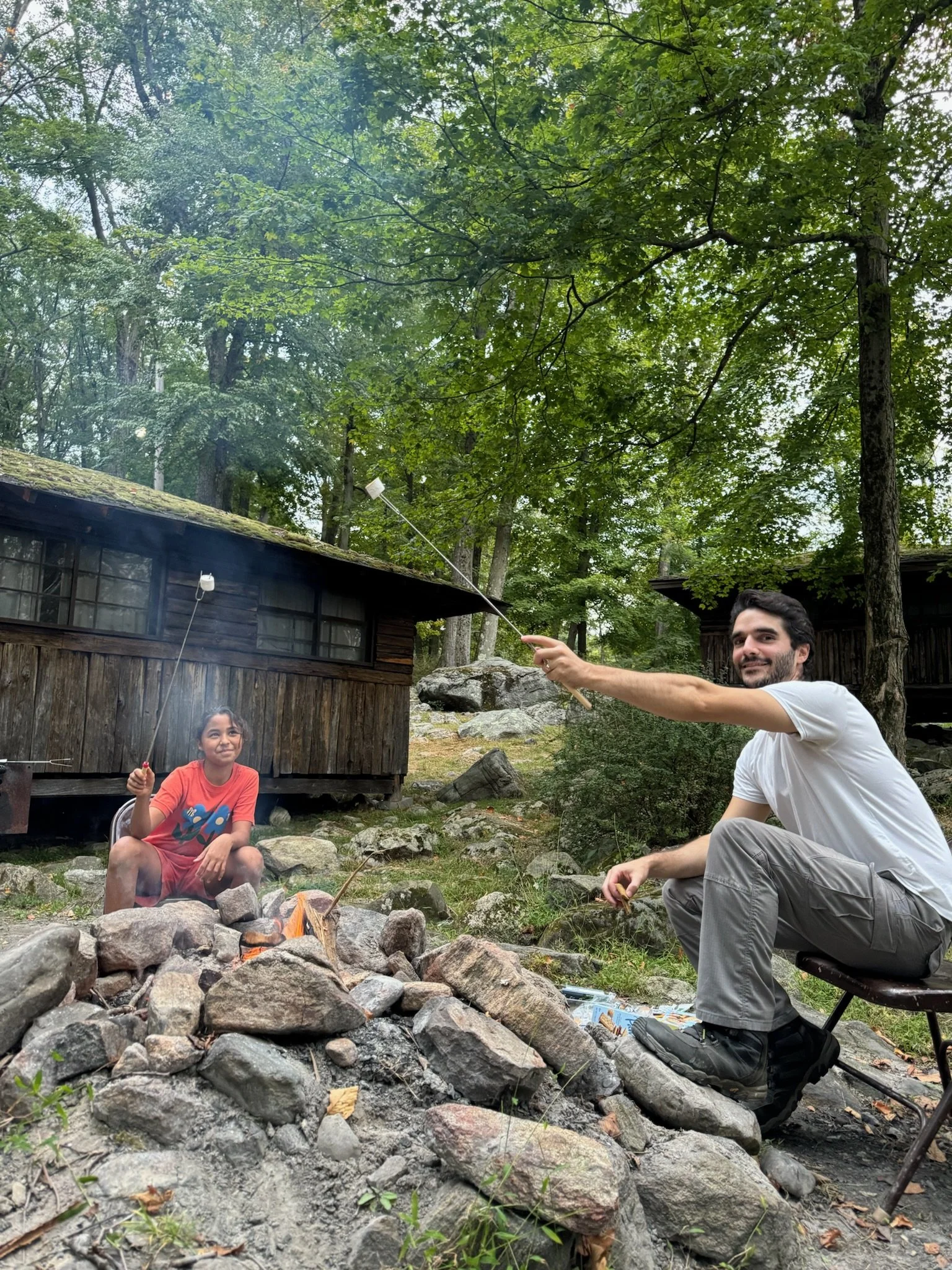 A young female student and a male teacher at Otto Specht School are sitting outside by two cabins, roasting marshmallows over a campfire built in the stones.