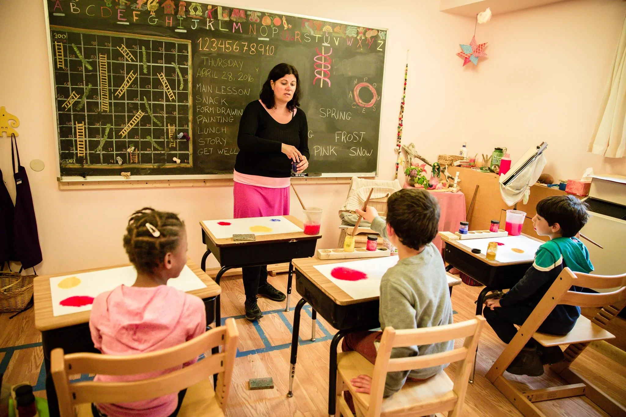 A female teacher at Otto Specht School stands in front of the blackboard teaching a group of young students who are working on watercolor paintings.