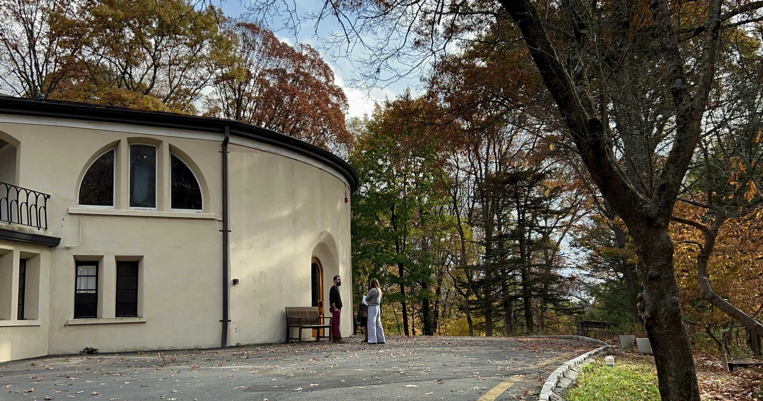 Two faculty members at Otto Specht School standing outside the main building on campus and talking on a Fall day, with colorful trees behind them.