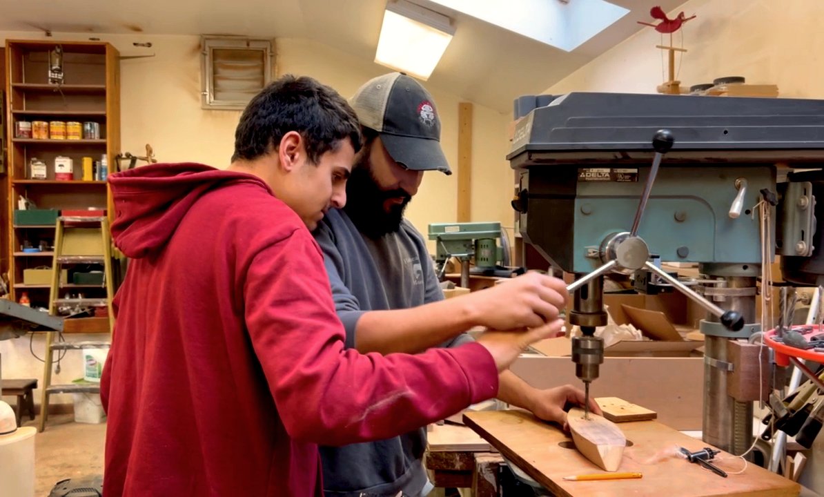 A male teacher and male high school student at Otto Specht School are working on a woodworking project together and operating a machine together over a piece of wood.
