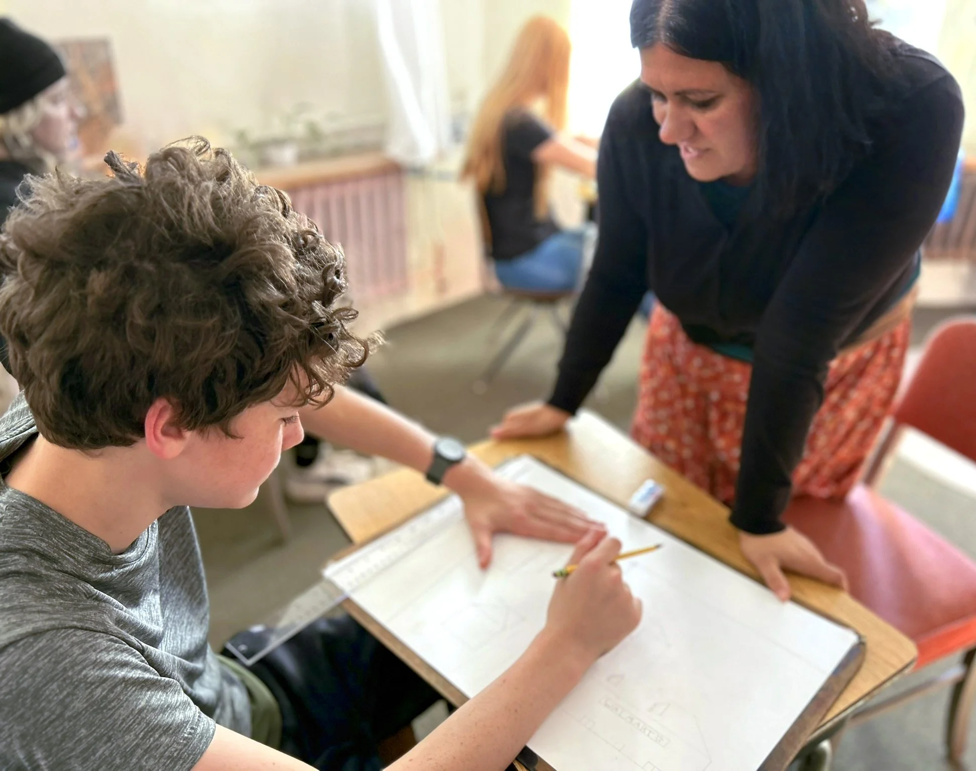 A female teacher leans over the desk of a male student to instruct him while he is working on his paper at Otto Specht School.