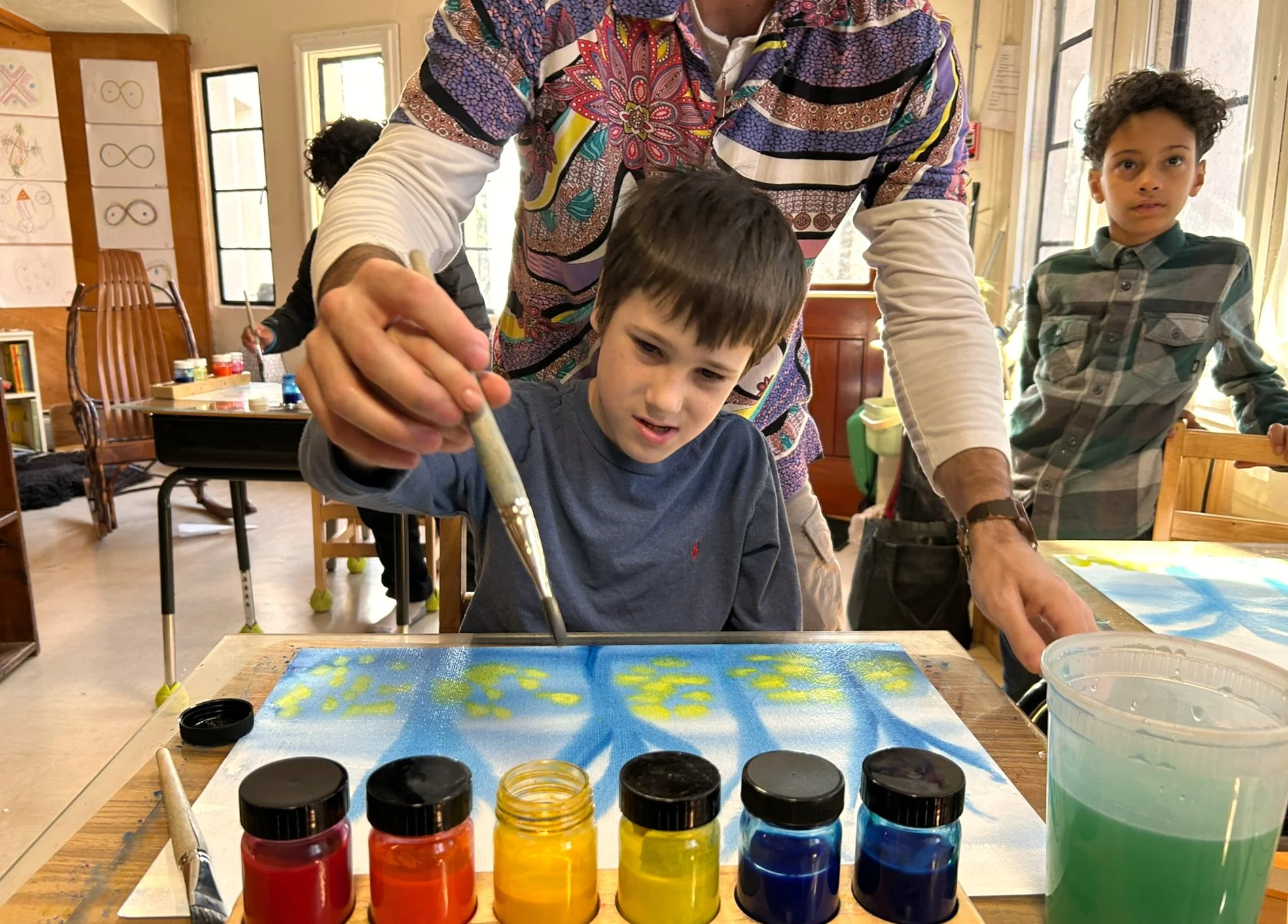 A teacher helps a young boy with his watercolor painting at Otto Specht School, while another young student boy looks on in the background. There is a row of rainbow watercolor paint pots on the desk in front of them.