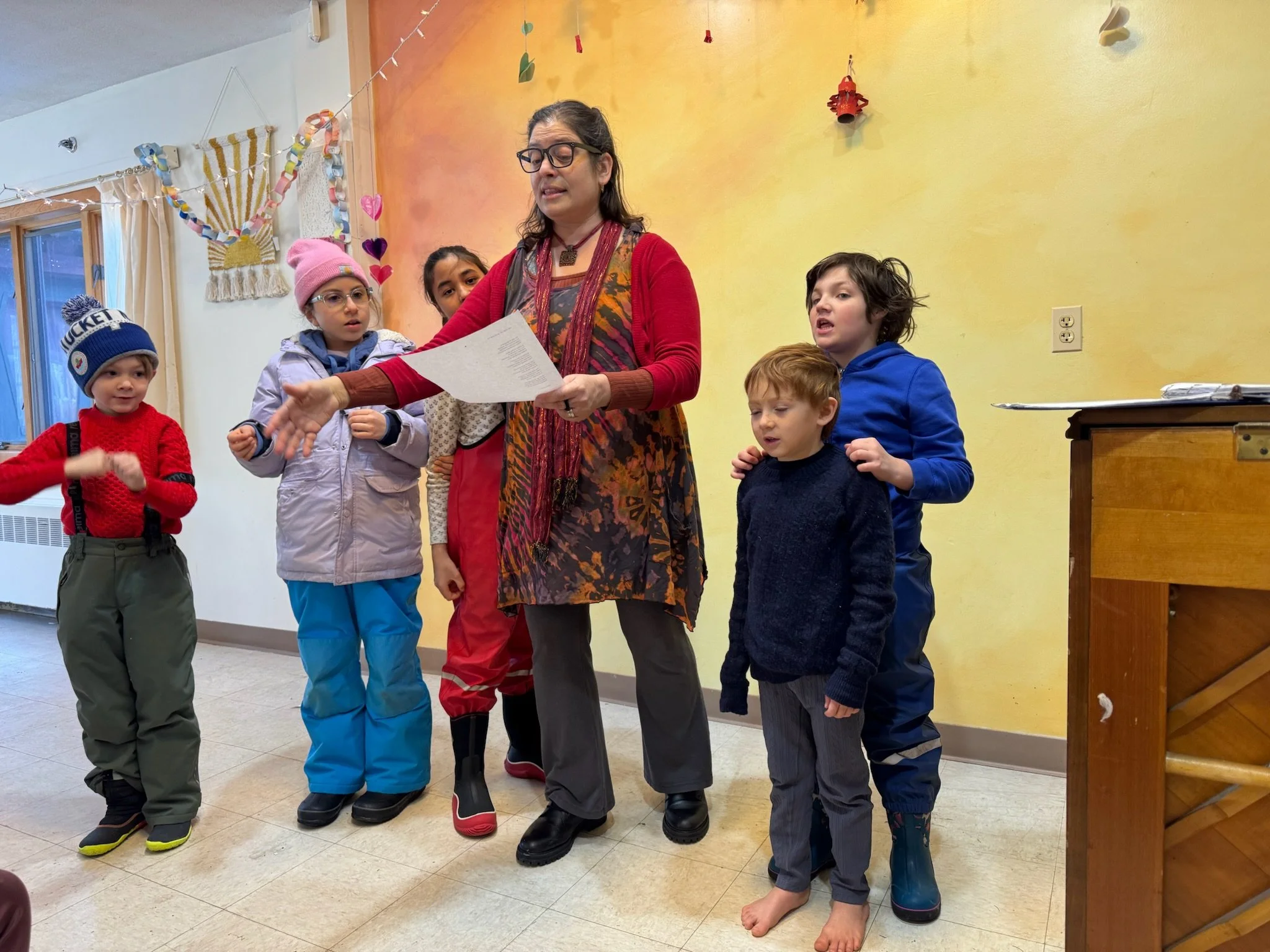 A teacher at Otto Specht School wearing a colorful red and dark purple outfit is reading something to the students who are surrounding her, who are all dressed in warm winter outdoor clothes. They are standing against an orange and yellow wall.