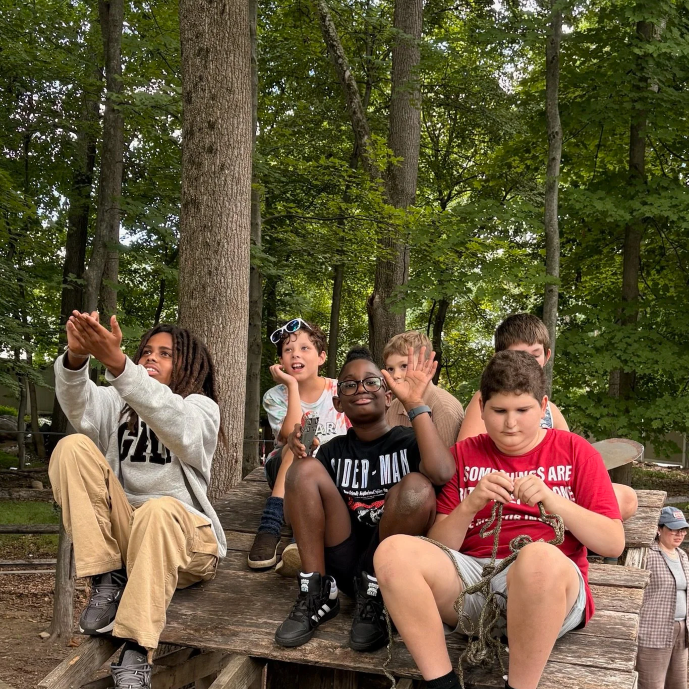 A group of boys of different ages and ethnicities at Otto Specht School are sitting on a wooden table outside, surrounded by green trees and smiling.