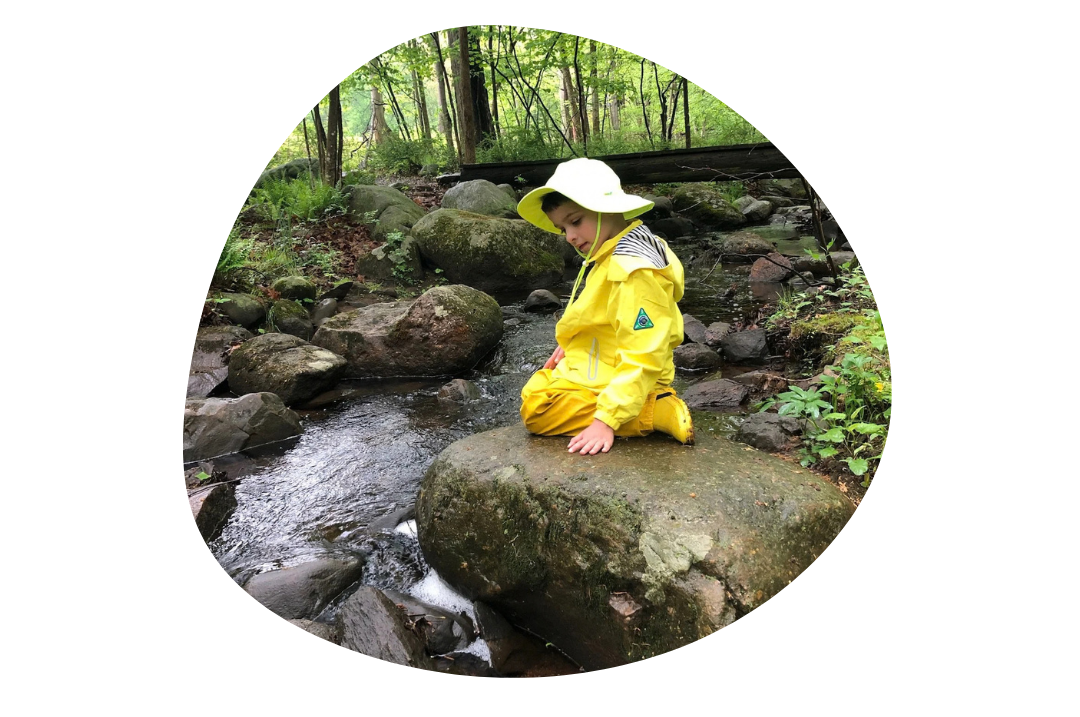 A kindergarten boy at Otto Specht School sits on a stone by a stream, wearing a yellow rain coat, boots, and hat.