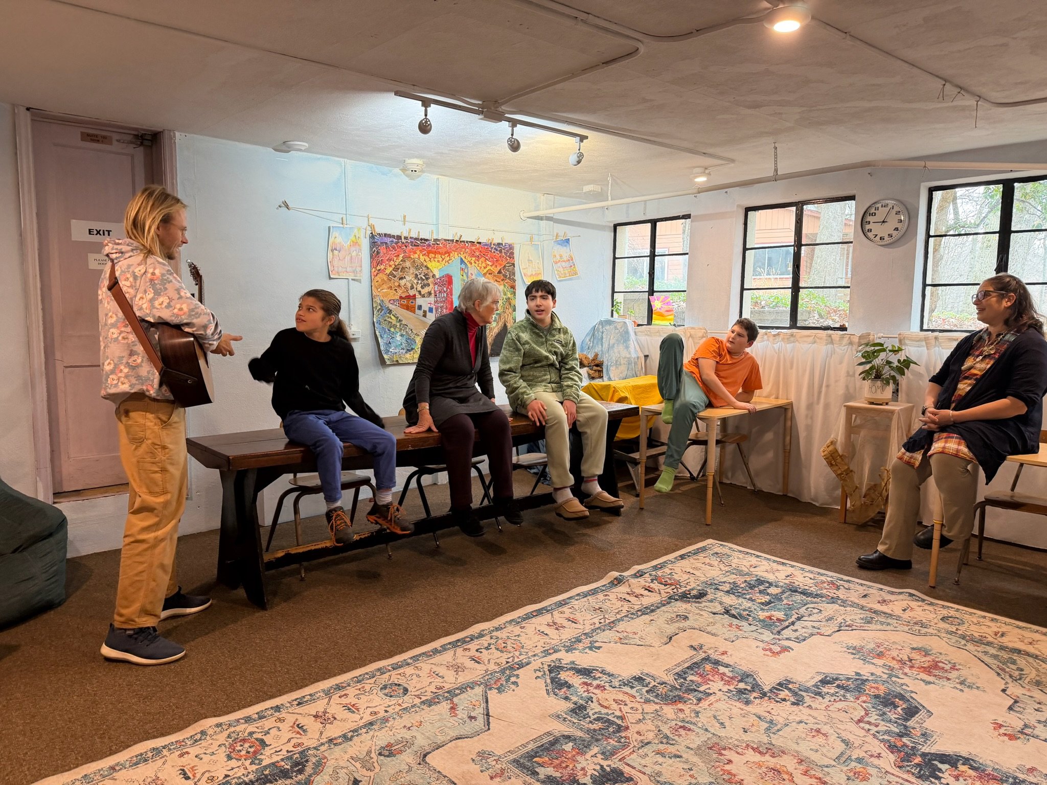A group of students and teachers gathered in a classroom at Otto Specht School. Some are standing and some are sitting on long tables at the edges of the room.