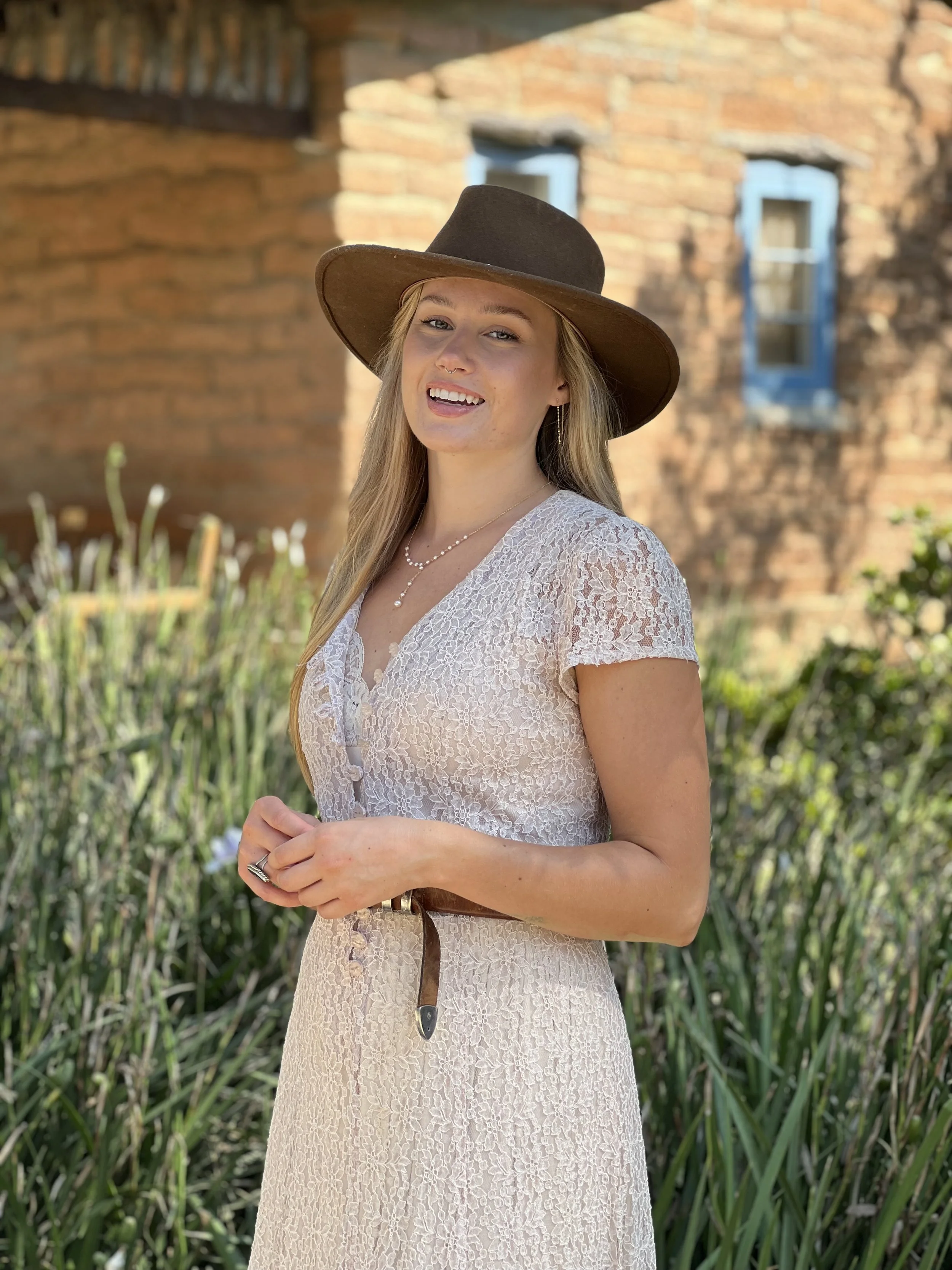 A woman with blonde hair wearing a wide-brimmed brown hat, a white lace dress, and jewelry, standing outdoors in front of a brick building with blue window frames and greenery.