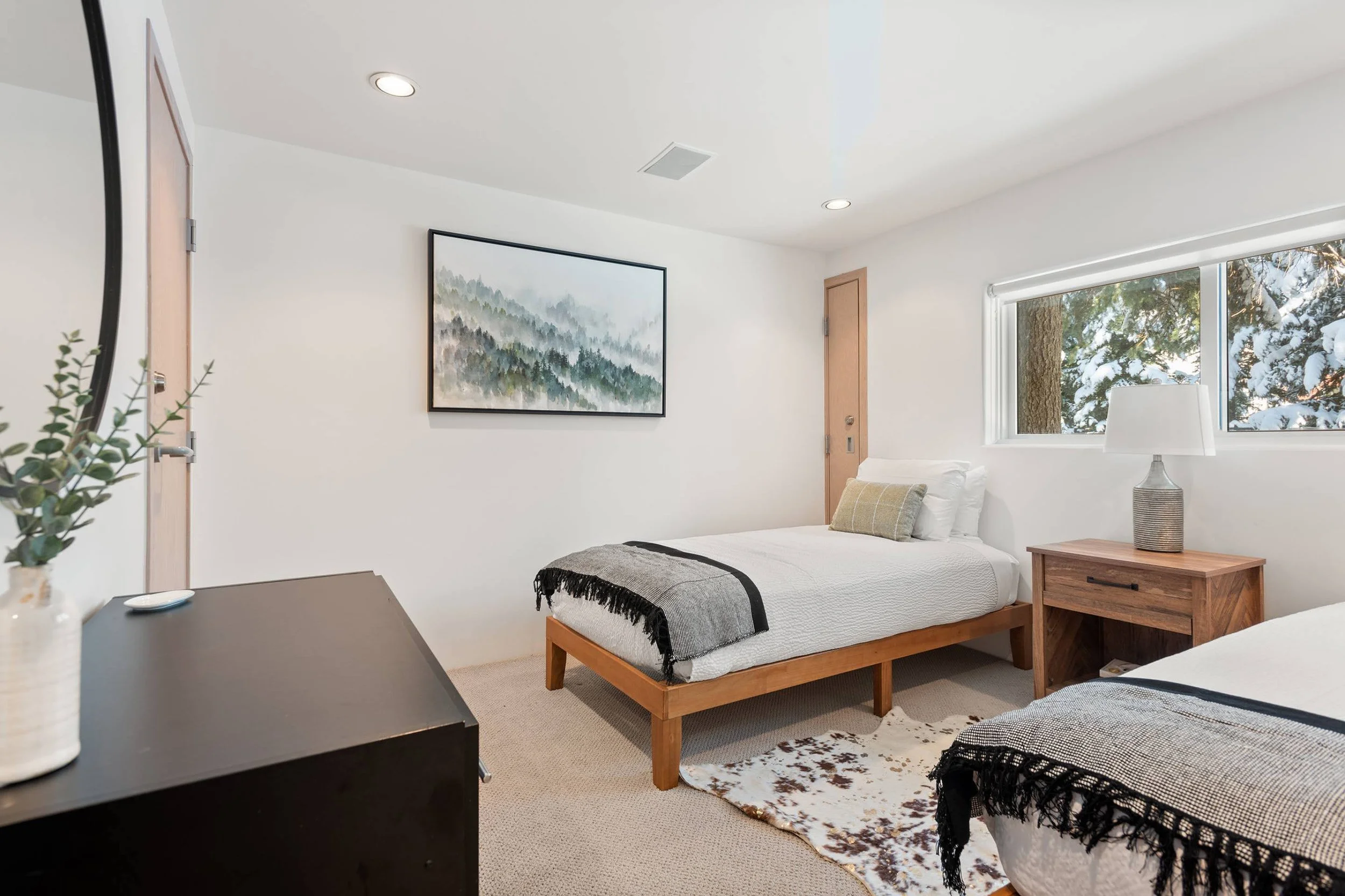 A small bedroom with a bed, a side table, and a window showing snow-covered trees outside. The room has white walls, a painting of a misty forest, and simple decor.
