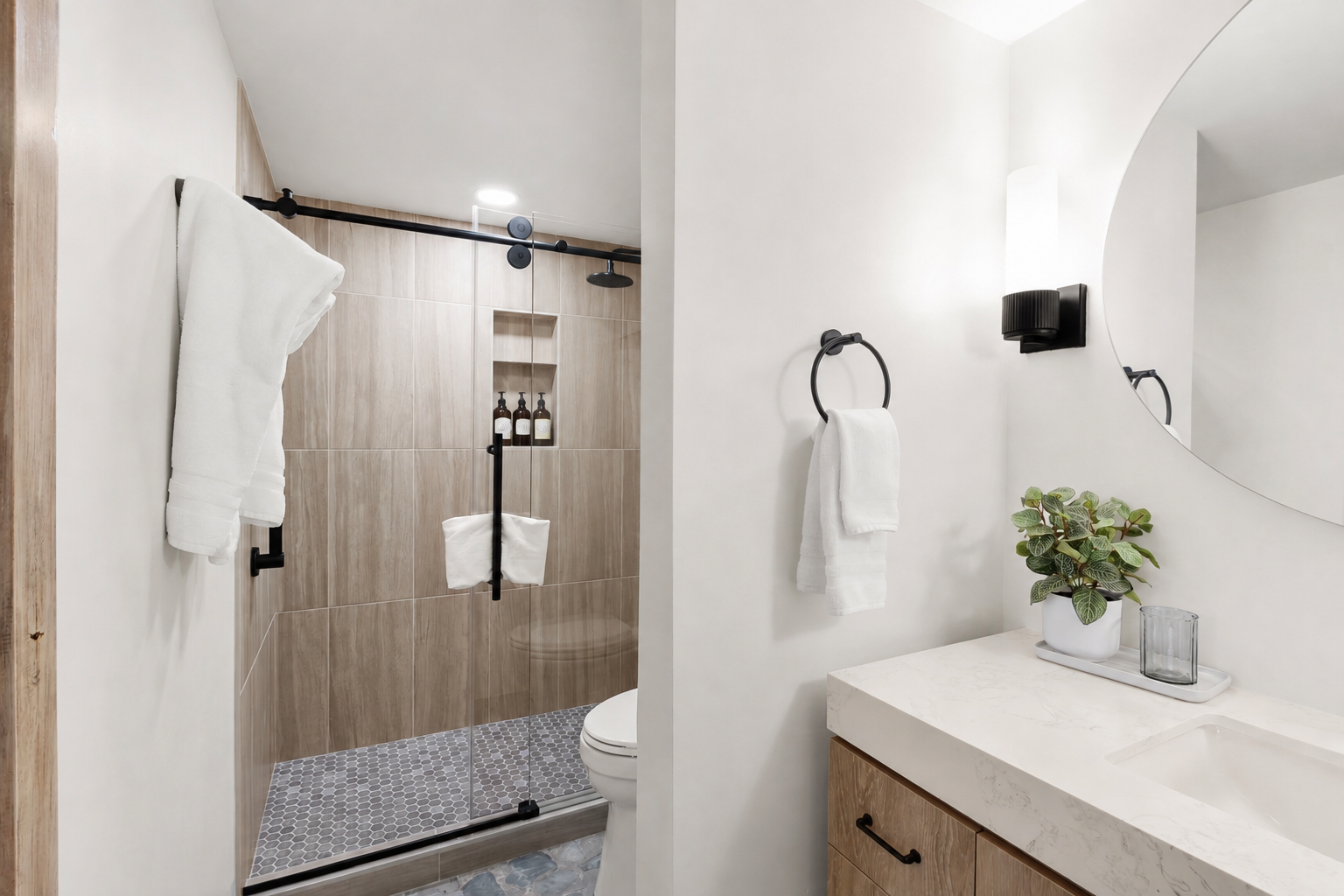 Modern bathroom with a walk-in shower with black hardware, a white toilet, a black towel ring with a white towel, a white vanity with a wooden cabinet, a large round mirror, a wall sconce, a potted plant, and a small tray with a glass container.
