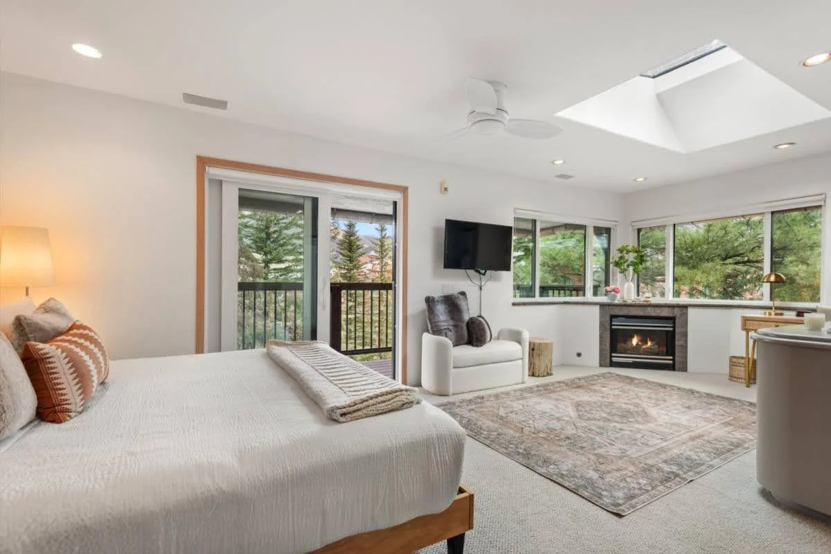 Modern bedroom with a white bed, pillows, throw blanket, sliding glass door leading to a balcony, a small loveseat, wall-mounted TV, fireplace, large windows with greenery outside, and a skylight.