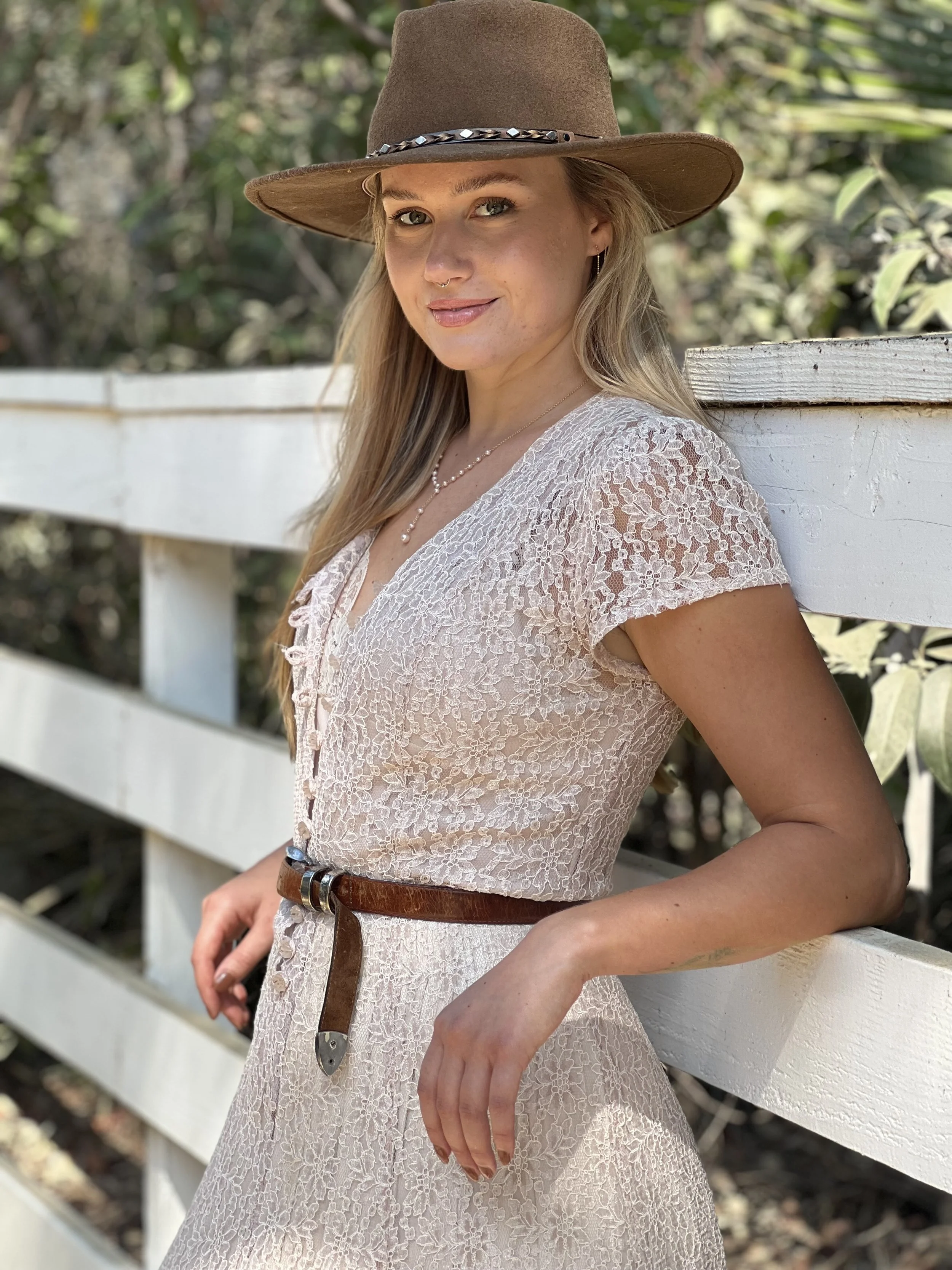 A young woman with blonde hair wearing a wide-brimmed brown hat and a white lace dress, standing outdoors near a white wooden fence with green foliage in the background.