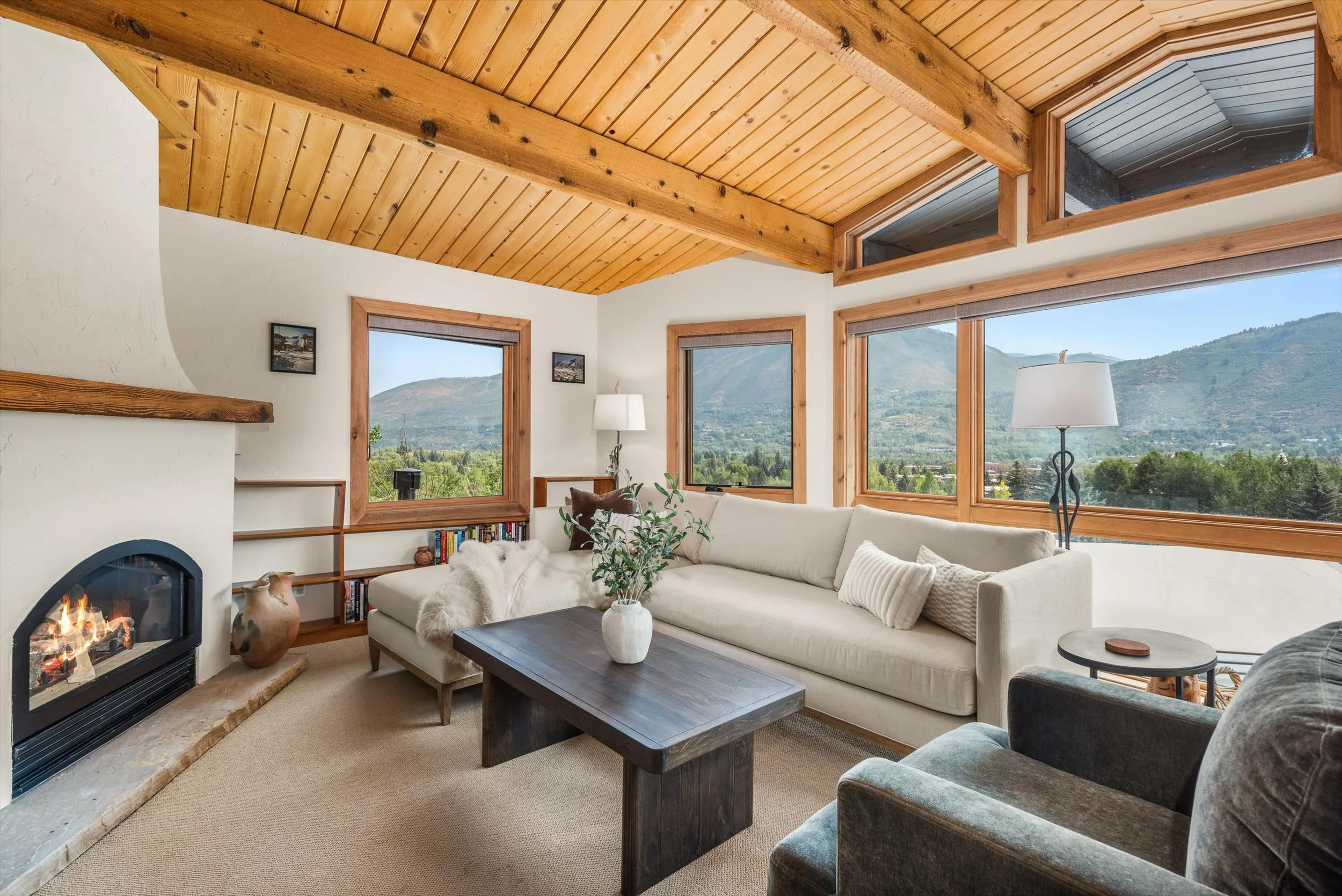 Living room with a white sectional sofa, black and gray armchair, wooden coffee table, fireplace, large windows showing mountain views, wood-paneled vaulted ceiling, ceiling fan, and decorative items.
