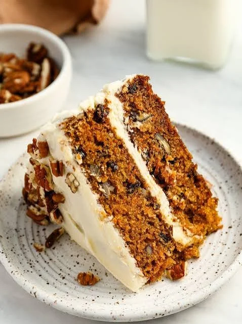Slice of carrot cake with cream cheese frosting and pecans on a white plate.