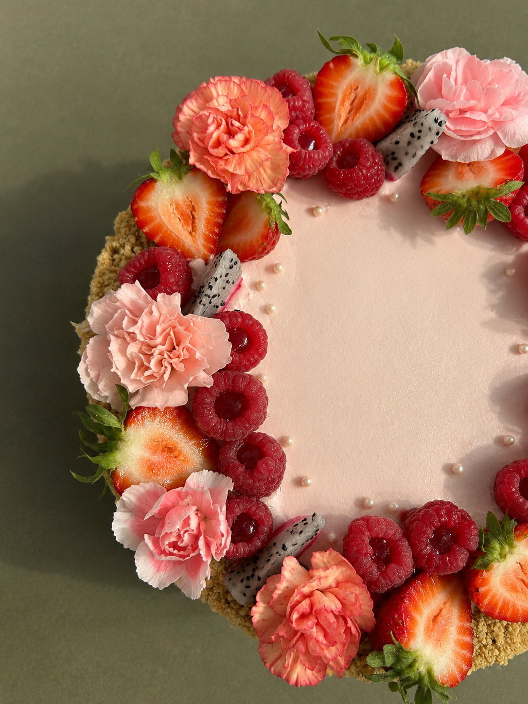 A close-up view of a decorated cake with pink frosting, topped with strawberries, raspberries, dragon fruit slices, pink carnations, and small edible pearls.