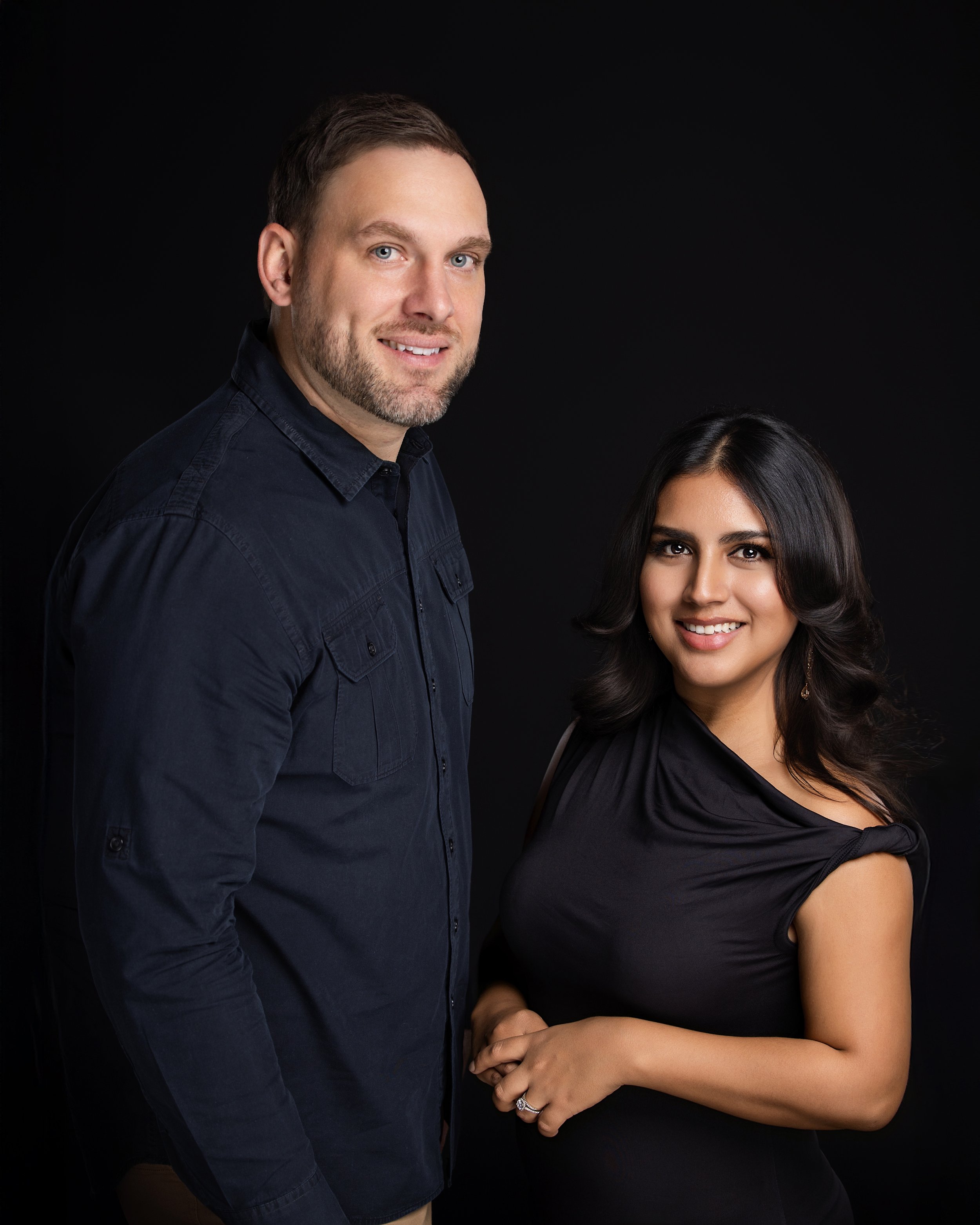 A man with short dark hair and a beard wearing a dark button-up shirt, standing next to a woman with long dark wavy hair and brown skin, wearing a black top with short sleeves, against a black background.
