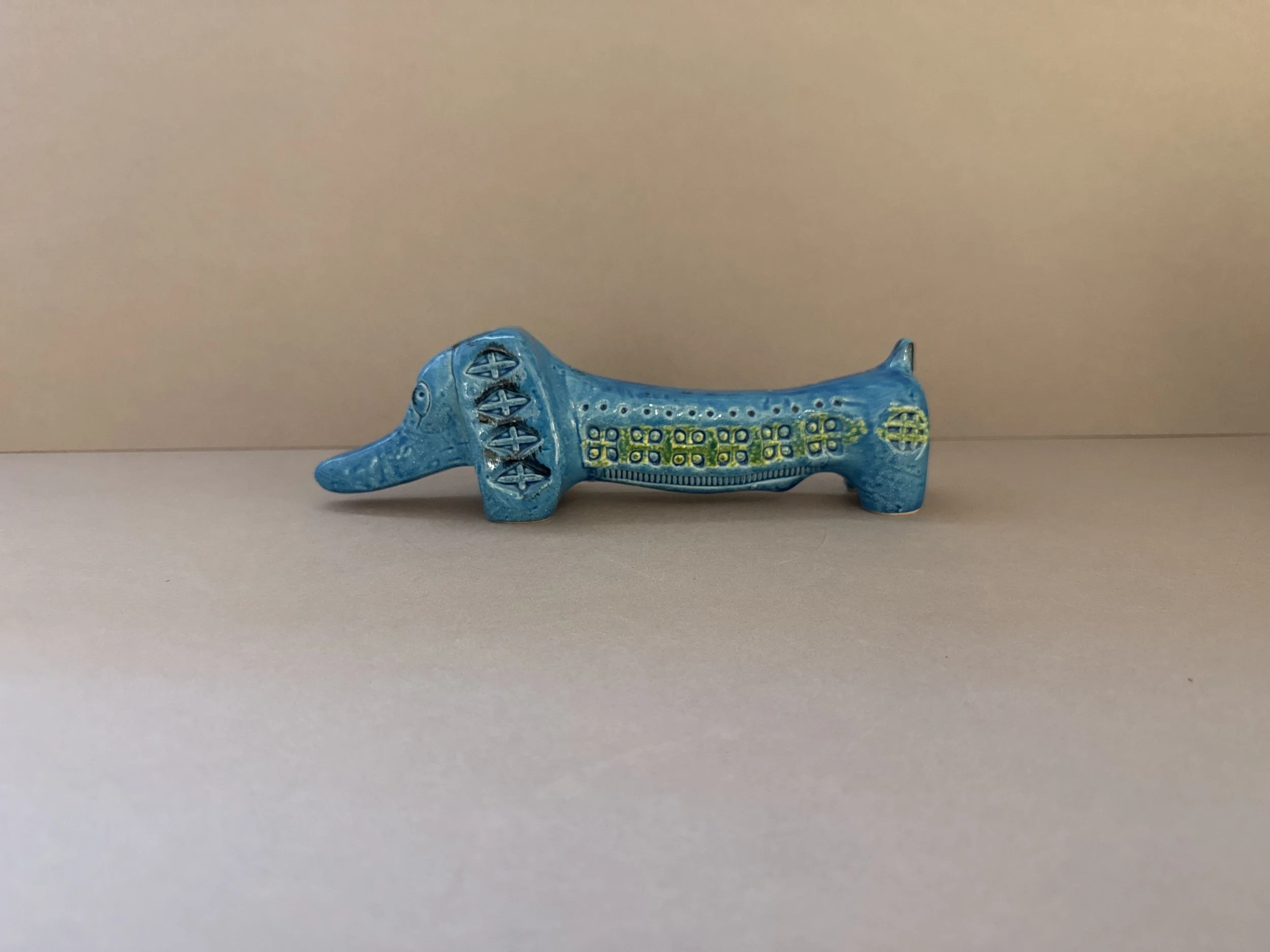 A small, blue, decorative ceramic or pottery animal figurine resembling a dog or similar creature with intricate patterns and designs on its body, placed on a plain, light-colored surface against a neutral background.