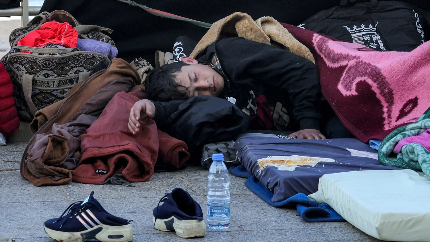 A young man sleeping on the street surrounded by blankets, clothes, and personal belongings, with a water bottle and sneakers in the foreground.