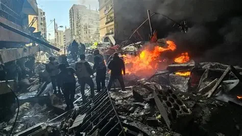 Scene of destruction caused by a fire with people standing amid debris and flames in a city street.