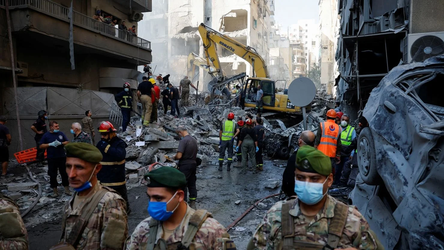 Emergency responders, soldiers, and workers at a building collapse site with rubble, debris, and damaged vehicles after an explosion or disaster.