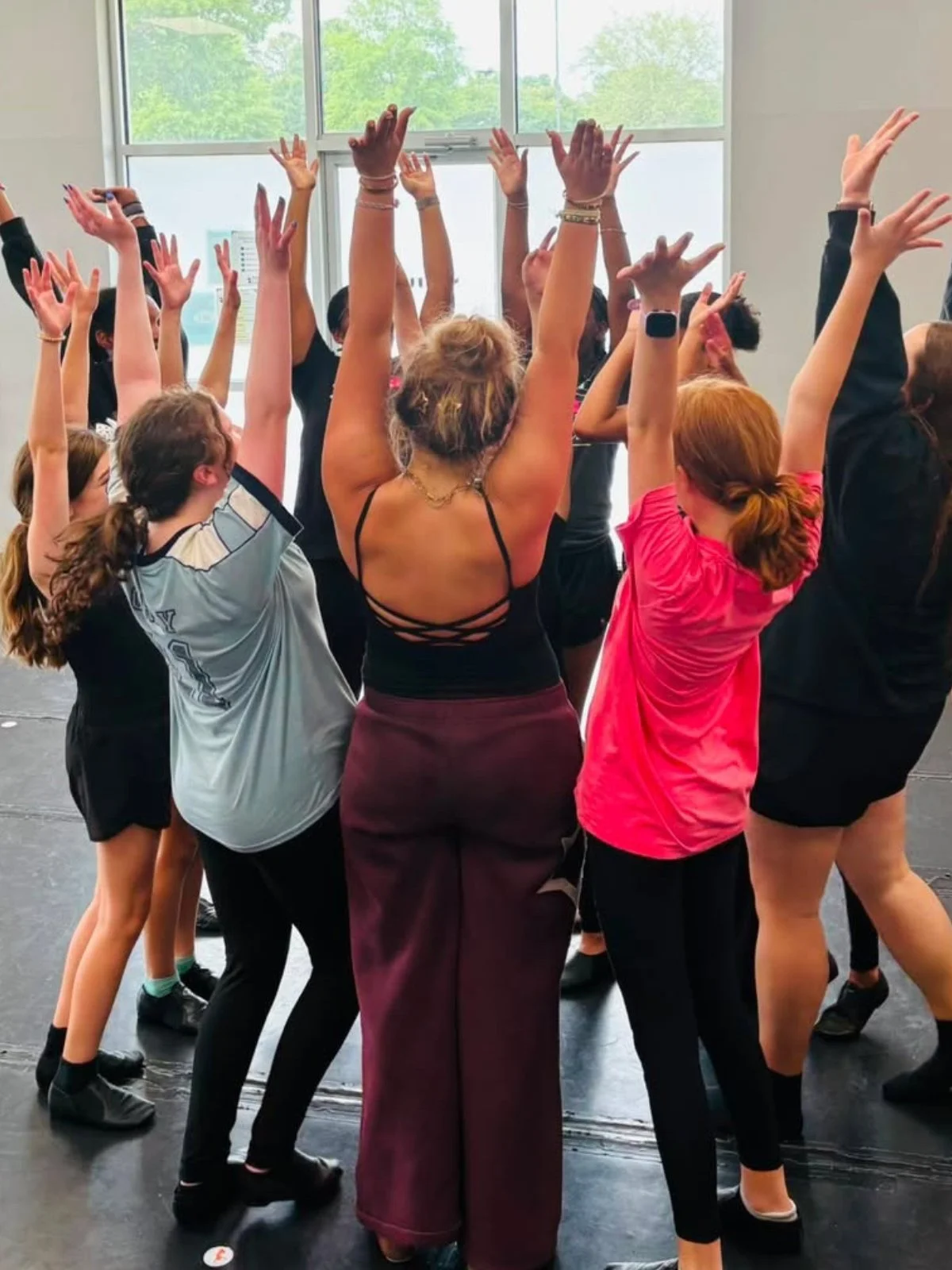 A group of children and a woman, with their arms raised, participating in a dance or exercise class indoors near large windows.