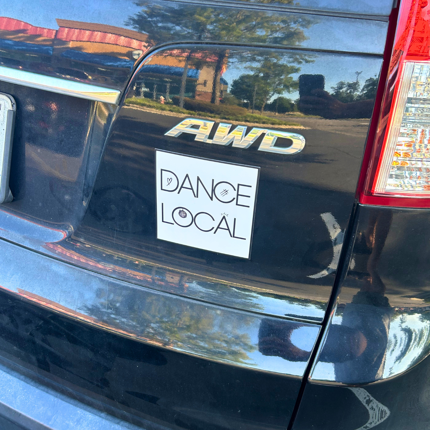 The rear of a dark-colored Honda Pilot vehicle with a sticker that says 'DANCE LOCAL' and includes icons of a heart, a hashtag, and a globe.