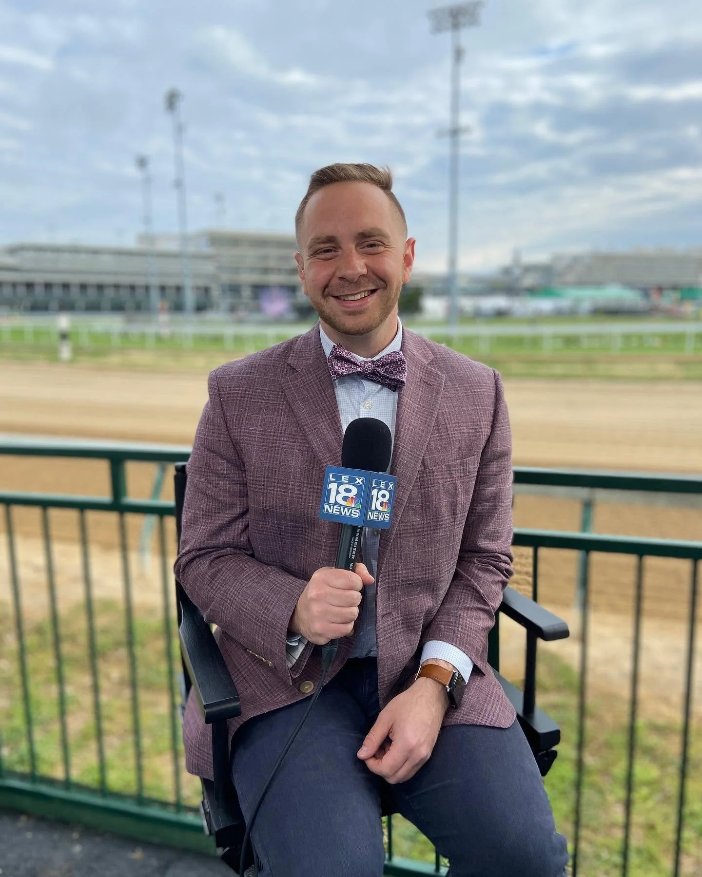 Austin Pollack in a plaid blazer and bow tie sitting outdoors, holding a microphone with the logo of a news station, in front of a racetrack with a grandstand and a cloudy sky.