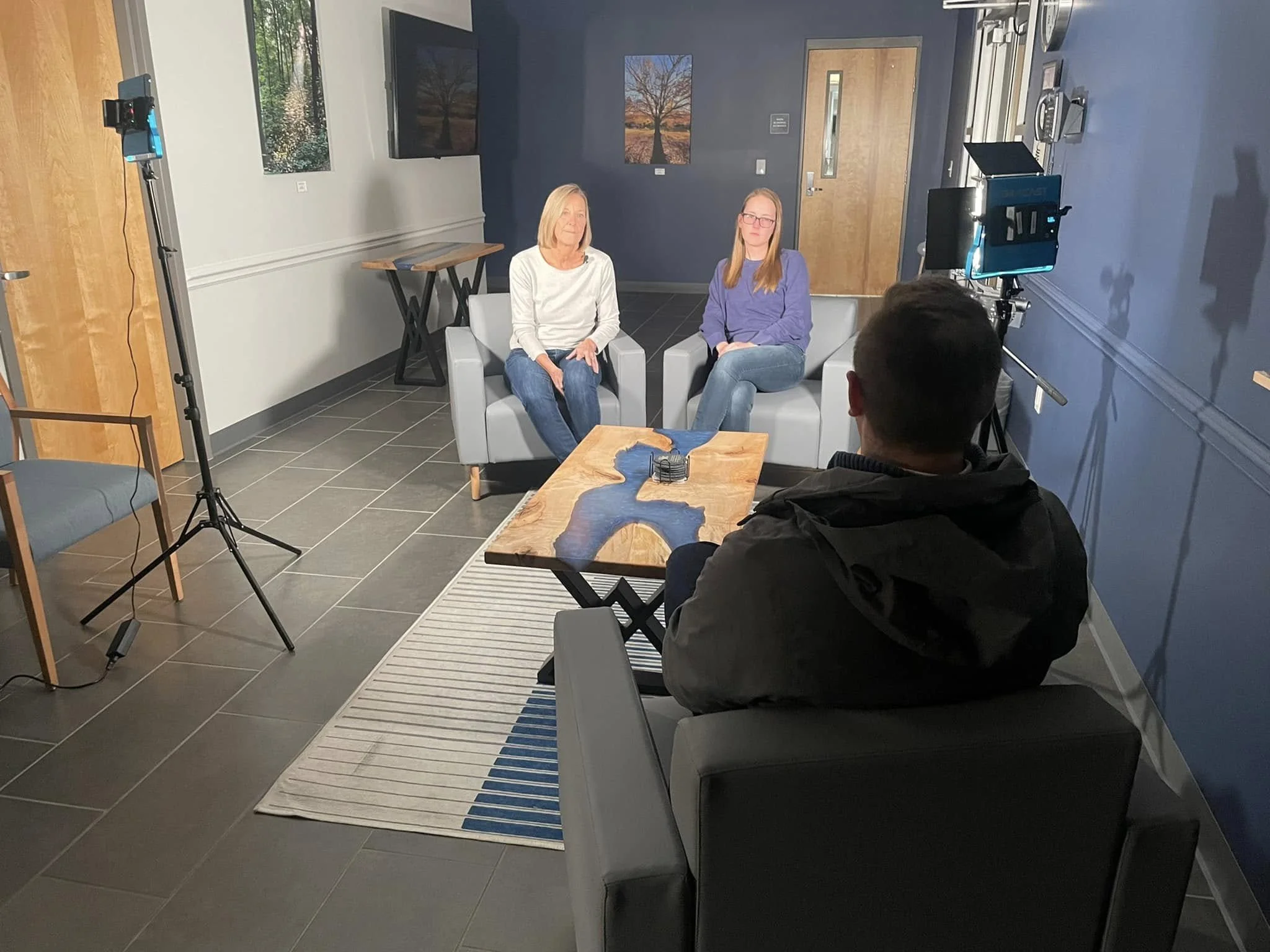 Two women are sitting on gray chairs in a room with dark blue walls, framed art, and a wood door. They are facing slightly towards Austin Pollack whose back is to the camera, with cameras and lighting equipment set up, filming an interview.
