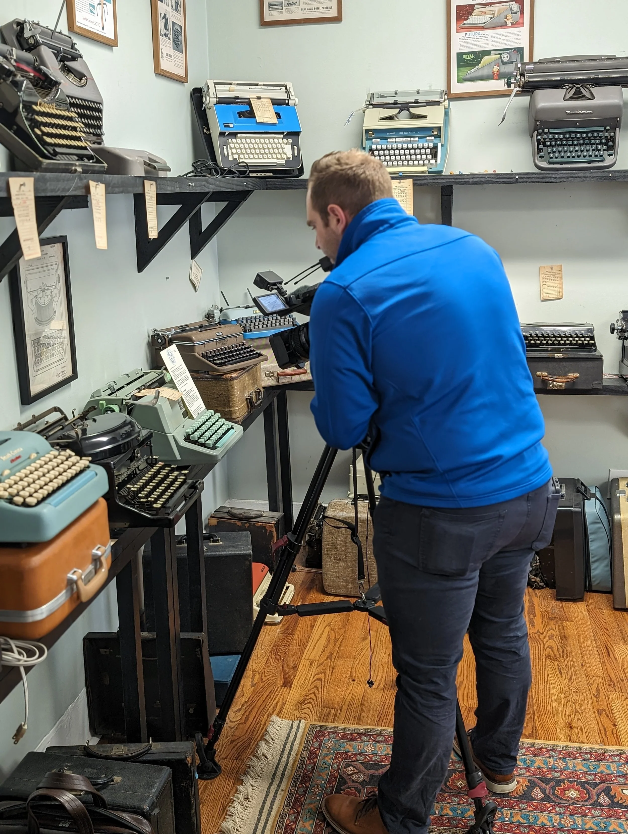 Austin Pollack in a blue jacket and dark jeans is filming with a video camera in a room filled with vintage typewriters displayed on black shelves. The room has hardwood floors and framed posters on the wall.