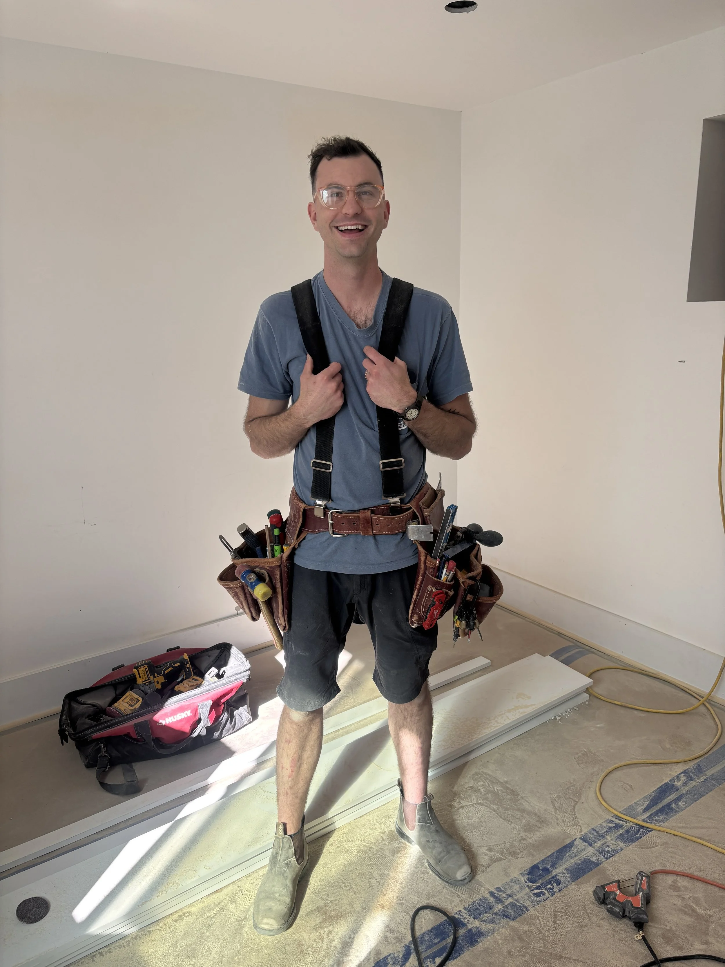 Photo of Max Holden standing in a room. He is wearing his tool harness and tool bags are on the ground behind him.
