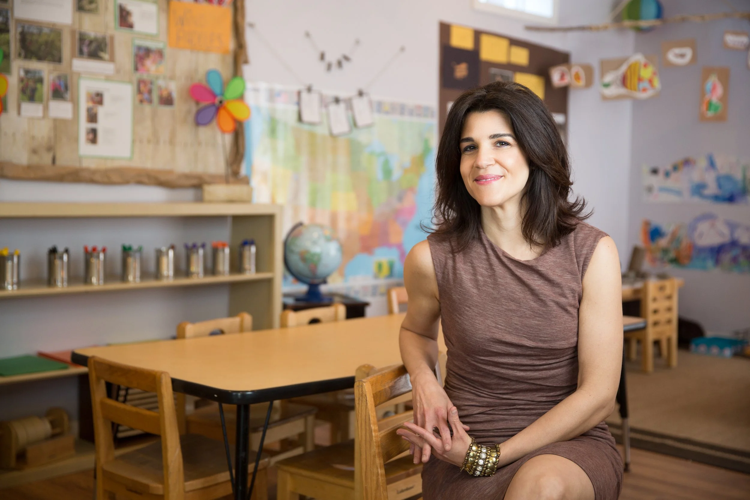 A woman with dark, wavy hair sitting in a classroom, smiling, with colorful educational decorations and maps on the wall behind her.