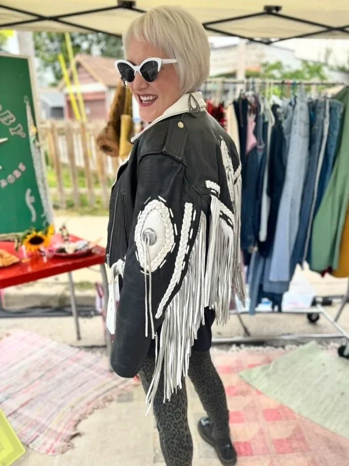 Woman wearing white sunglasses and a black leather jacket with white fringe details, standing at an outdoor market with clothing racks and a table with items in the background.