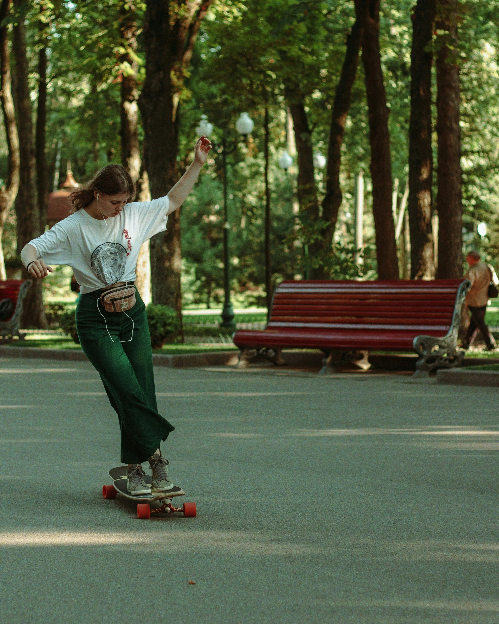 A young woman skateboarding on a paved path in a park with trees and benches in the background during daylight.