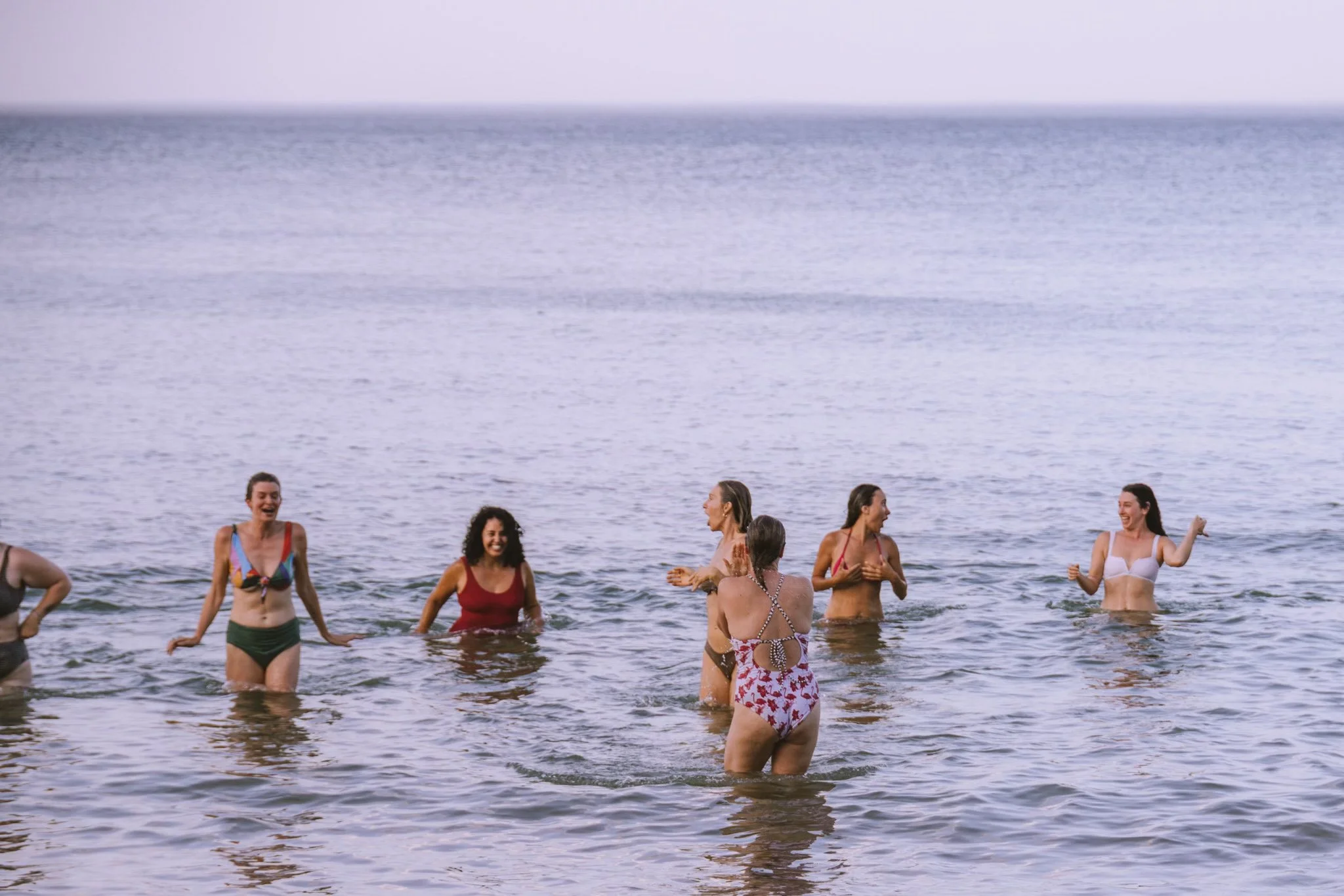 A group of women in swimsuits playing and enjoying themselves in the water at the beach with an expansive ocean in the background.