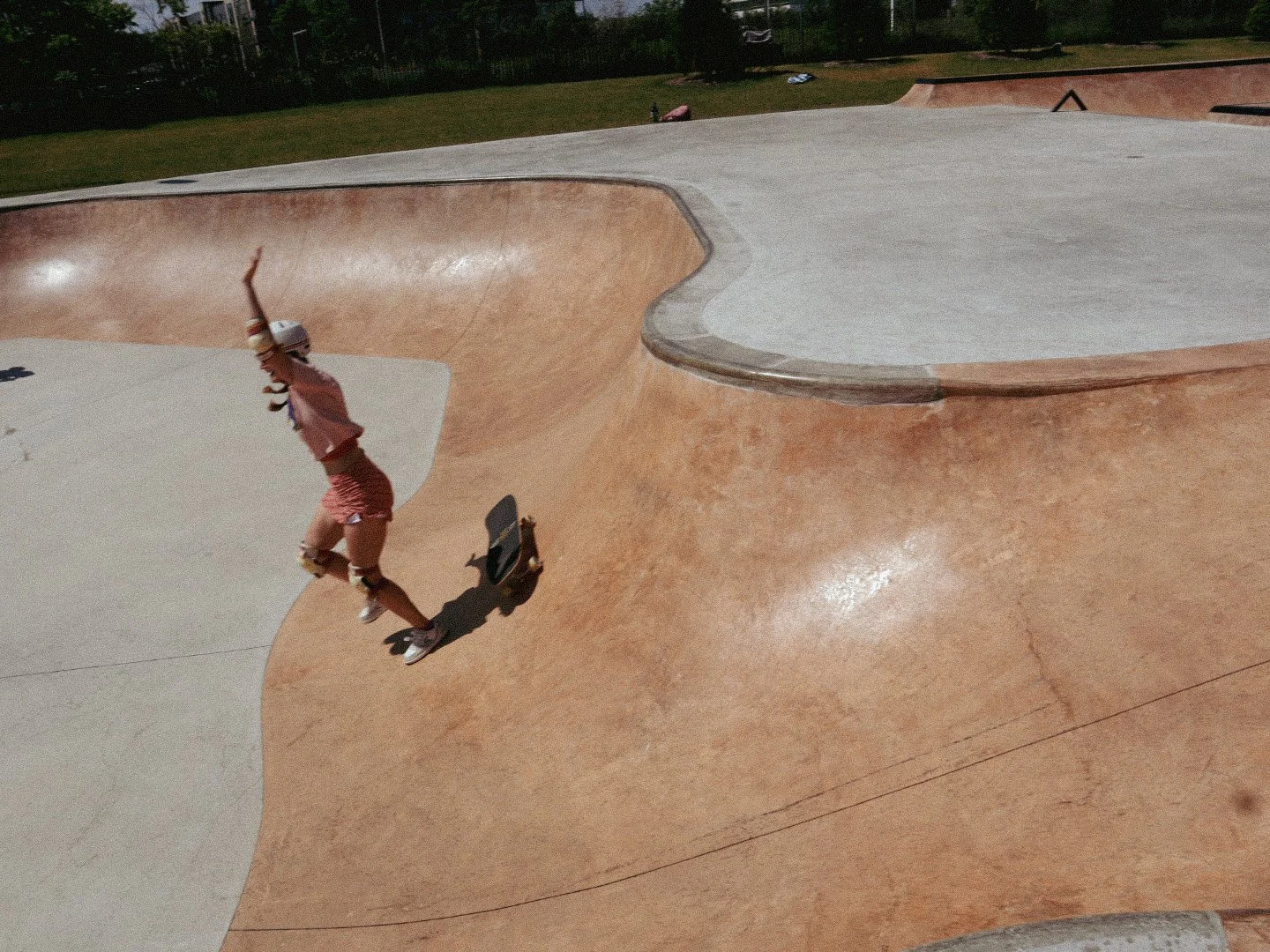 A young girl wearing a pink hat and matching outfit skating in a concrete skatepark with curved ramps and bowls, surrounded by green trees and grass.