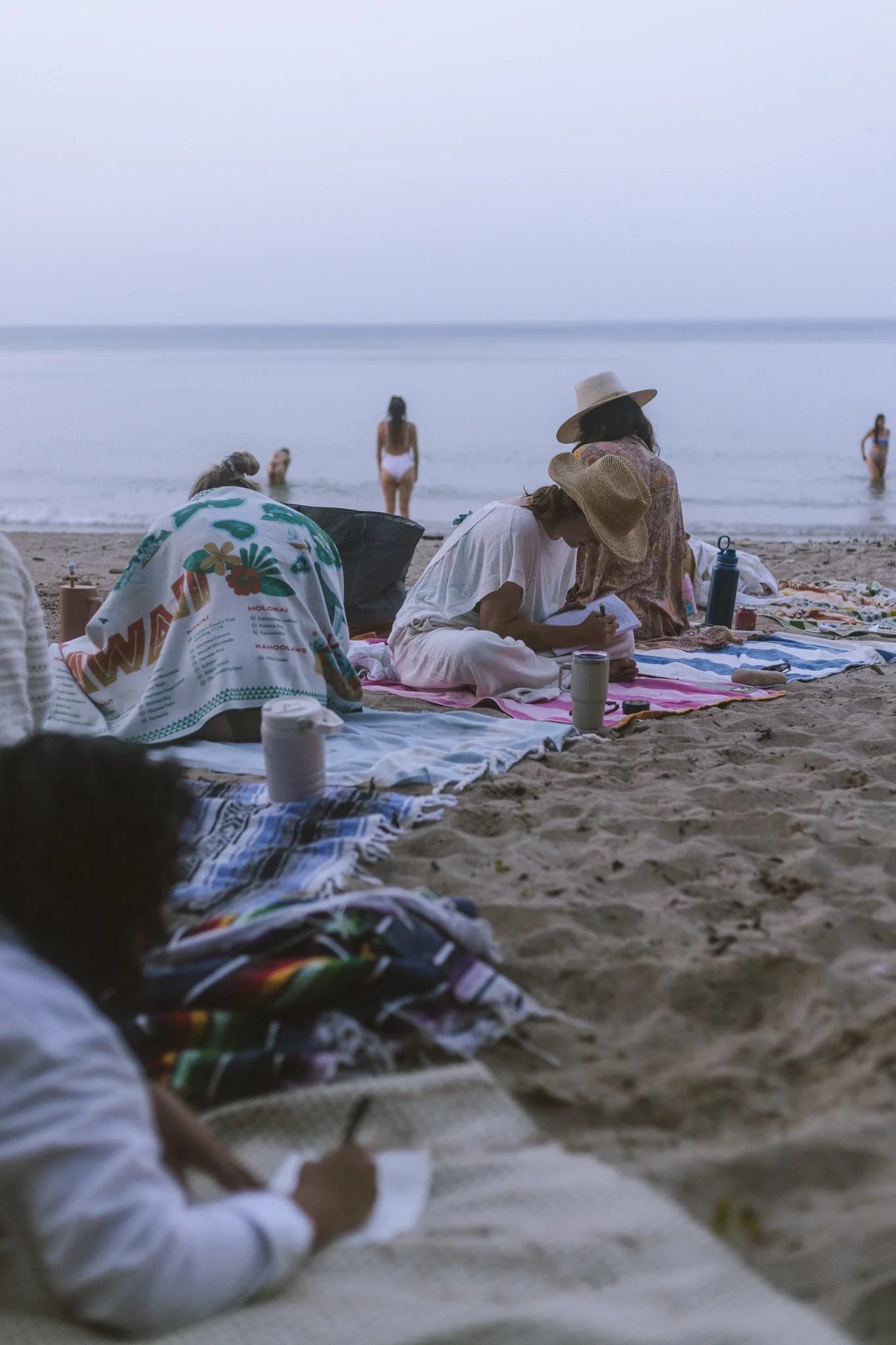 People sitting on a beach with towels, some writing or drawing in notebooks, with others swimming or standing at the water's edge in the background.