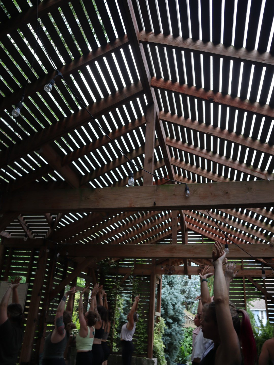 Group of people participating in an outdoor yoga or stretching session under a wooden pergola with string lights.