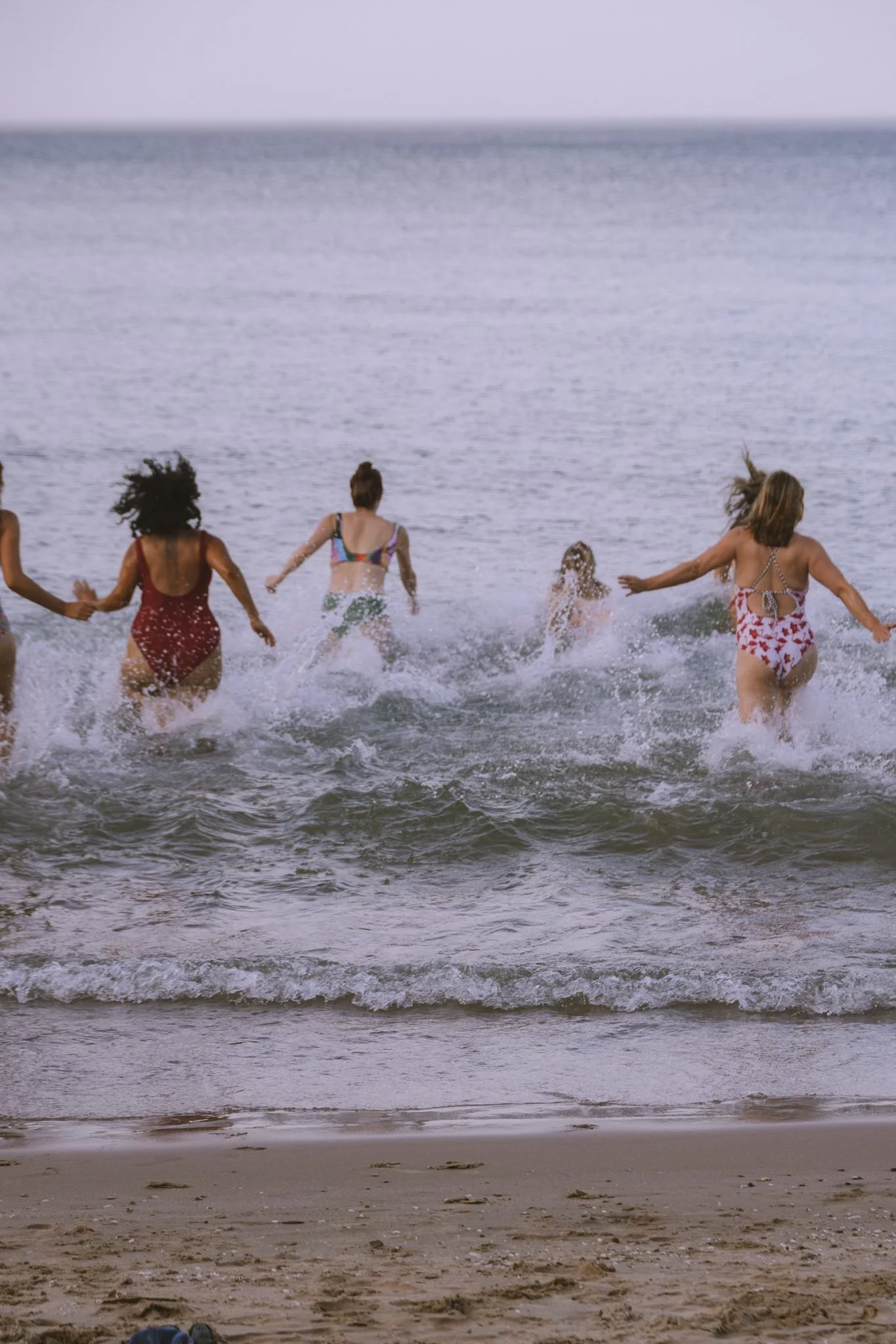 Several women running into the ocean at the beach, wearing swimsuits, with waves and the sky in the background.