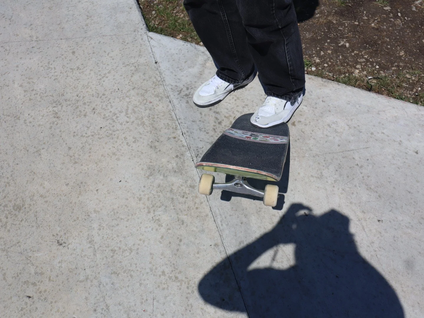 Person standing on a skateboard on a concrete sidewalk, wearing white sneakers and black jeans, with part of their shadow visible on the ground.