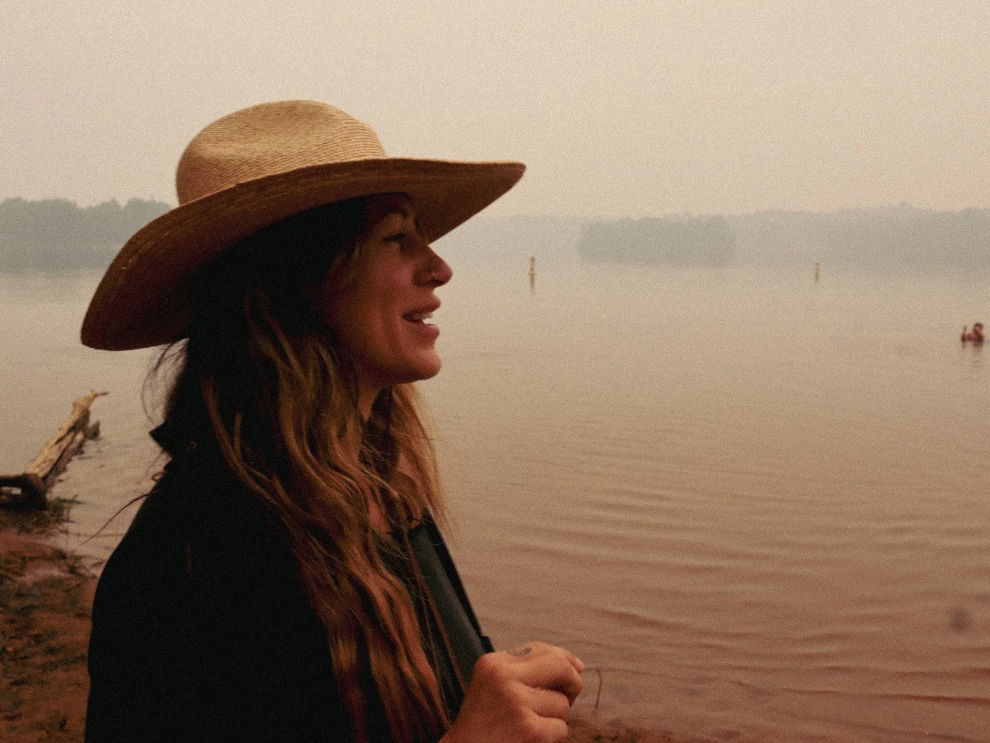 A woman with long wavy hair wearing a large straw hat and dark clothing, standing on a beach at sunset, smiling and looking towards the water while holding a stick.