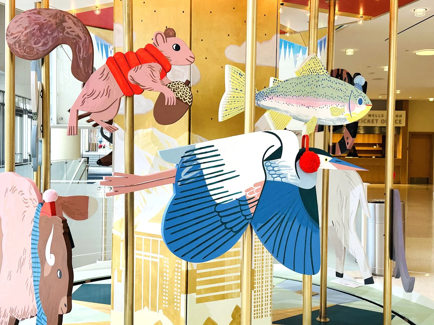 image of a blue heron with ear muffs, a squirrel in a puffer vest, a rainbow trout with a beanie, and a bison with a beanie as animals on a carousel