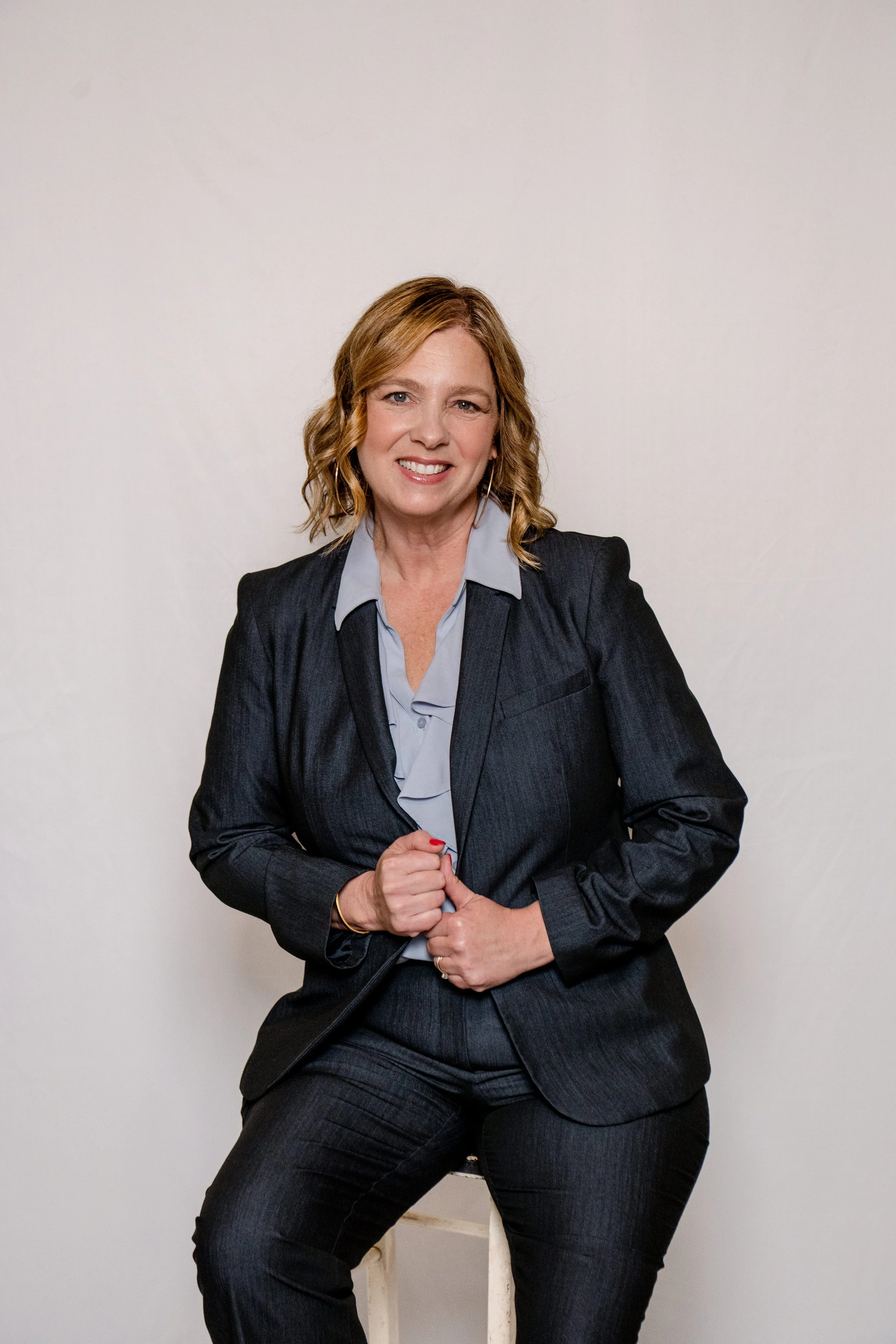 A woman with shoulder-length wavy blonde hair wearing a dark business suit with a light blue shirt, sitting on a white stool against a plain light background, smiling and looking at the camera.