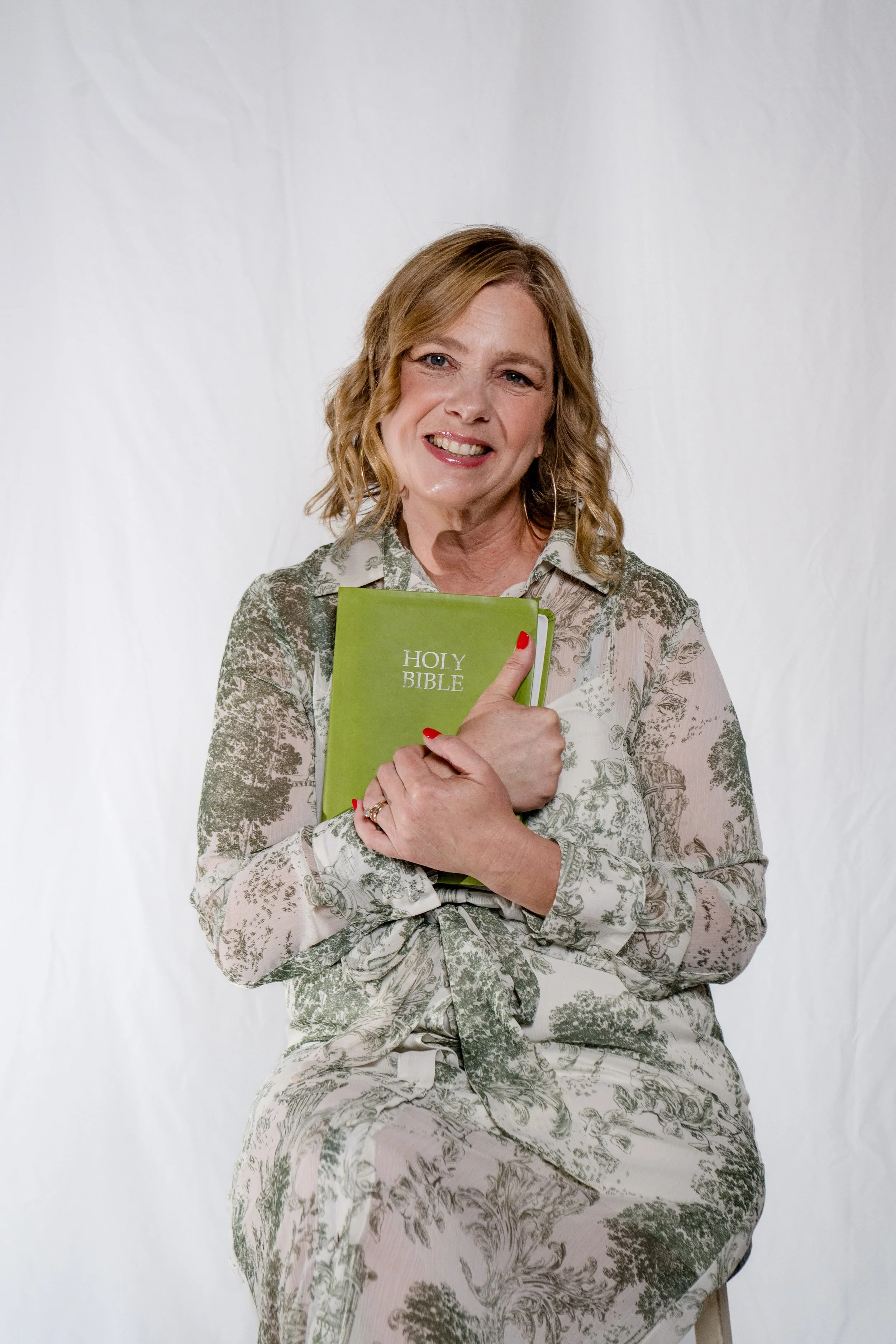 A woman with shoulder-length curly hair smiling and holding a green Holy Bible in front of her chest, sitting against a plain white background.